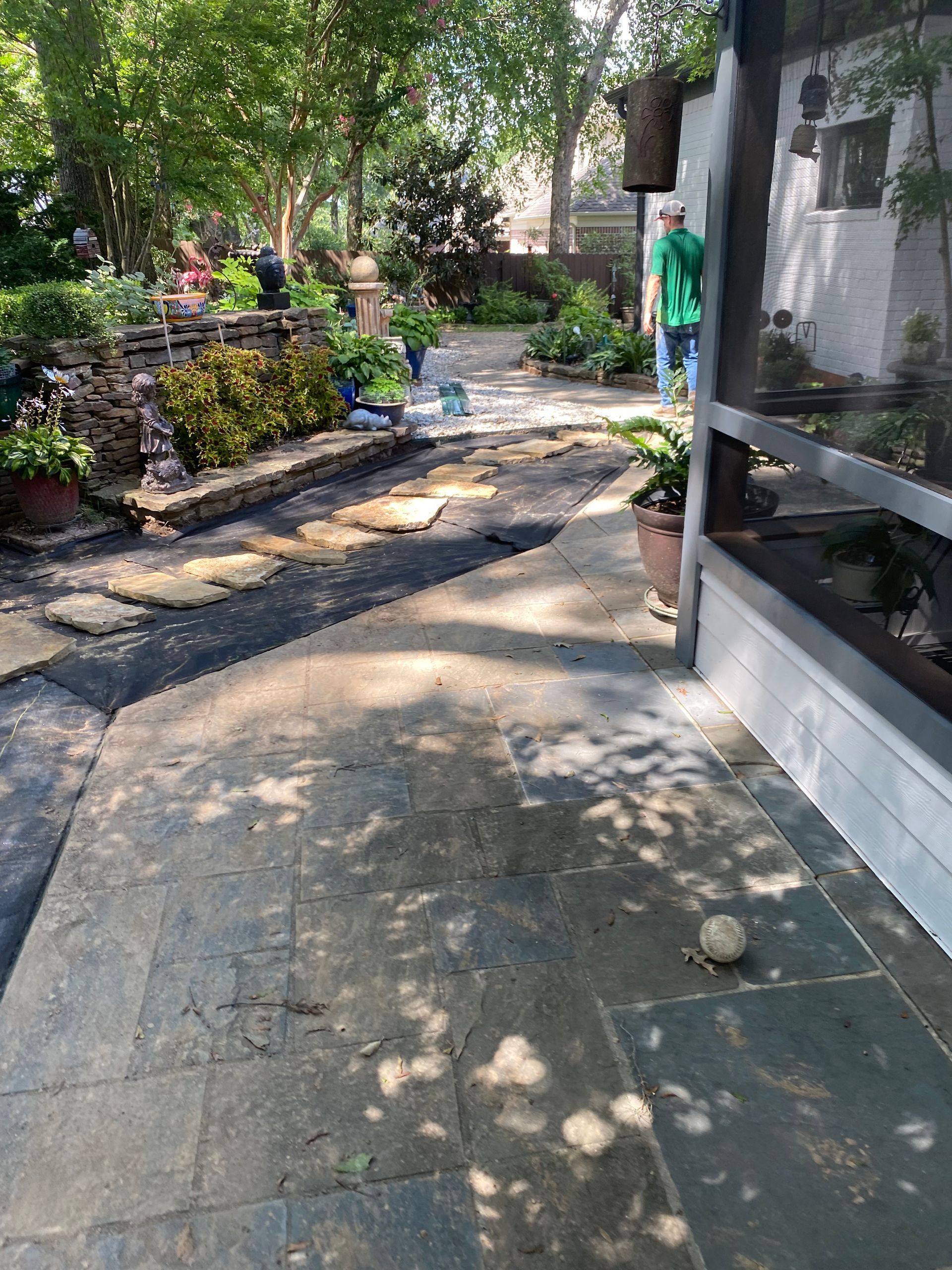 Patio with flagstone, leading to a garden with plants and a person in green. Shadows from overhead trees are cast on the ground.