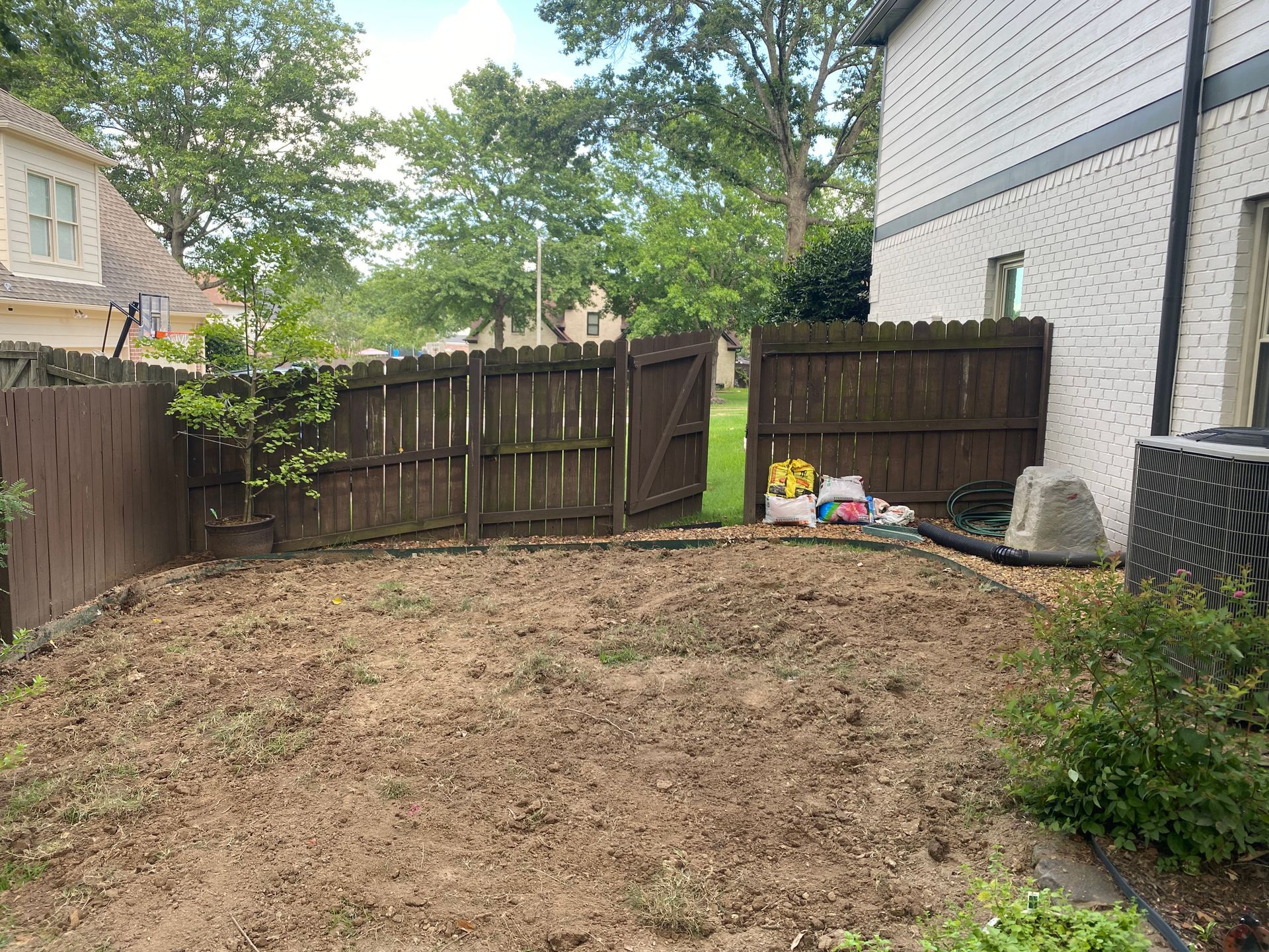 A backyard with a dirt patch, brown wooden fence, and a white brick house. Some debris lies near the fence.