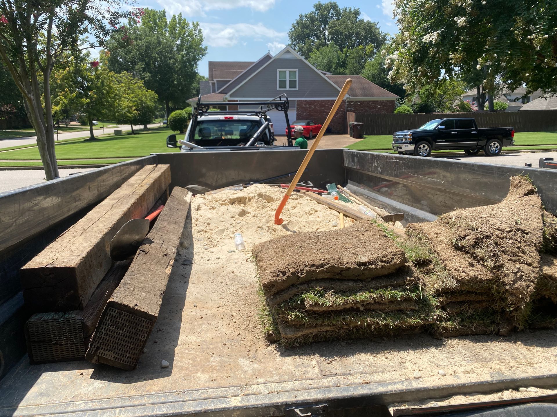 Truck bed filled with landscaping materials: wooden beams, soil, and sod on a sunny day in a residential area.