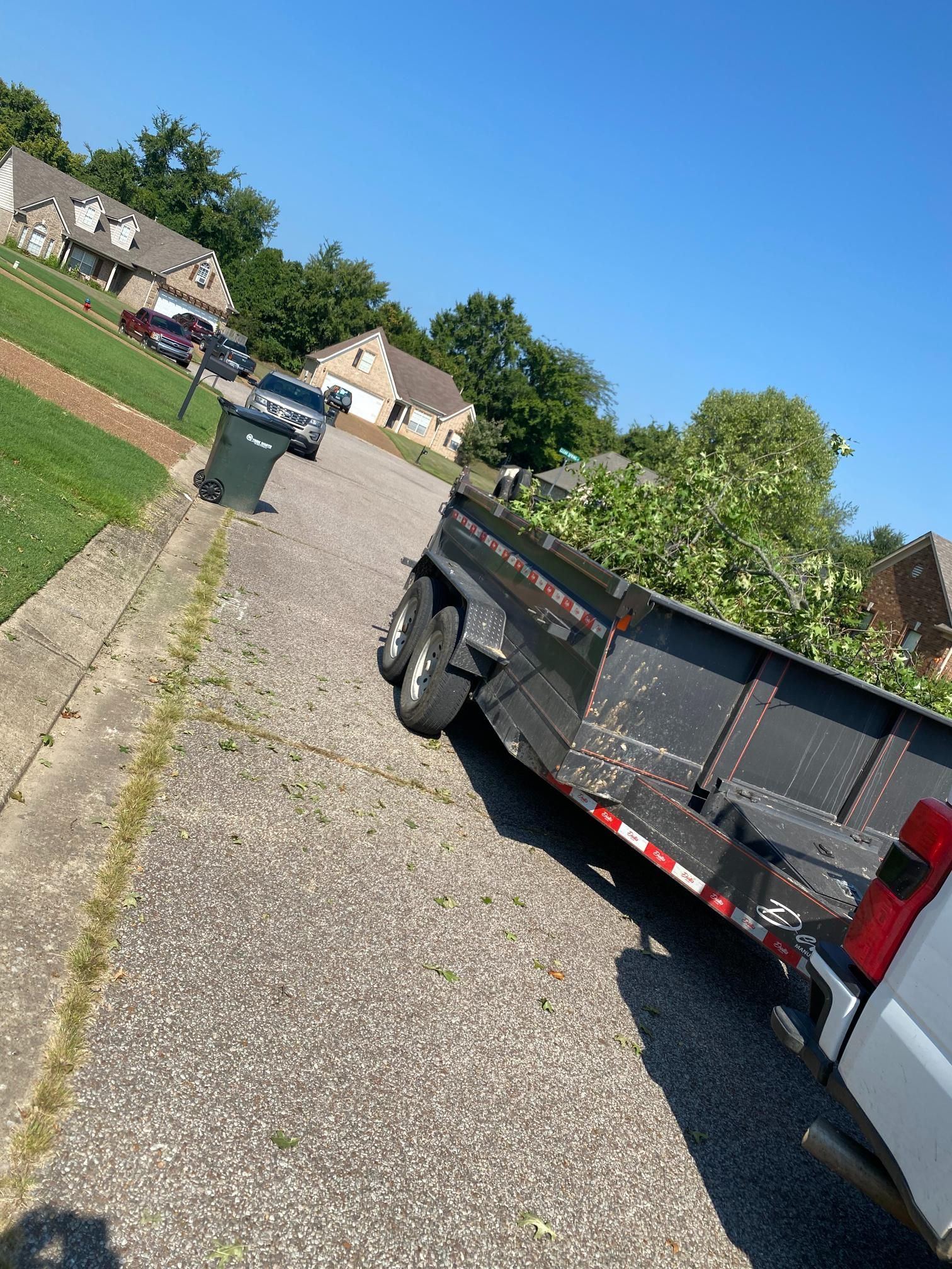 A black trailer loaded with green yard waste is parked on a street with a white truck attached. Houses line the background.