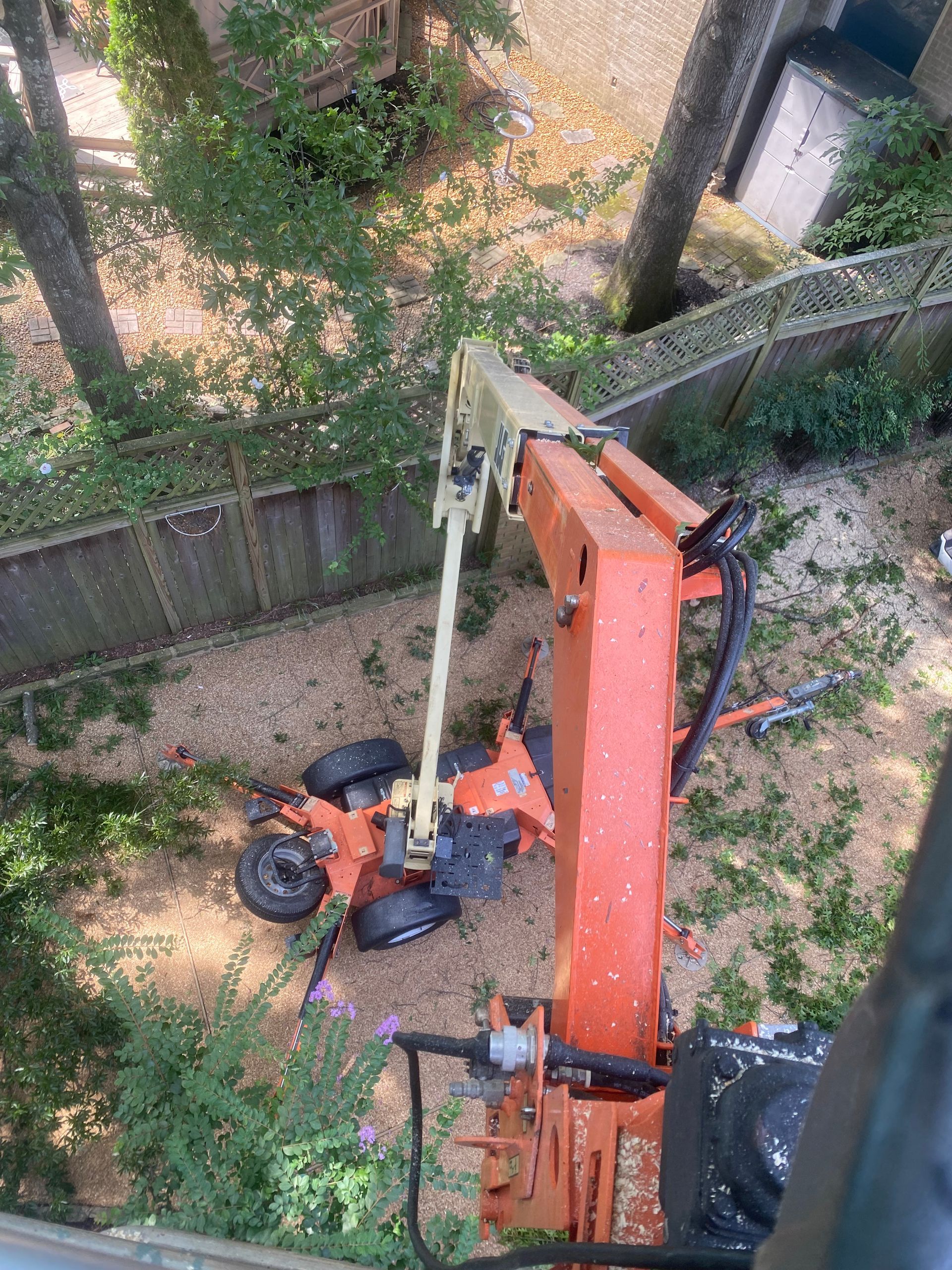 A crane operating near trees and a wooden fence. The crane is orange, and the scene is outdoors.