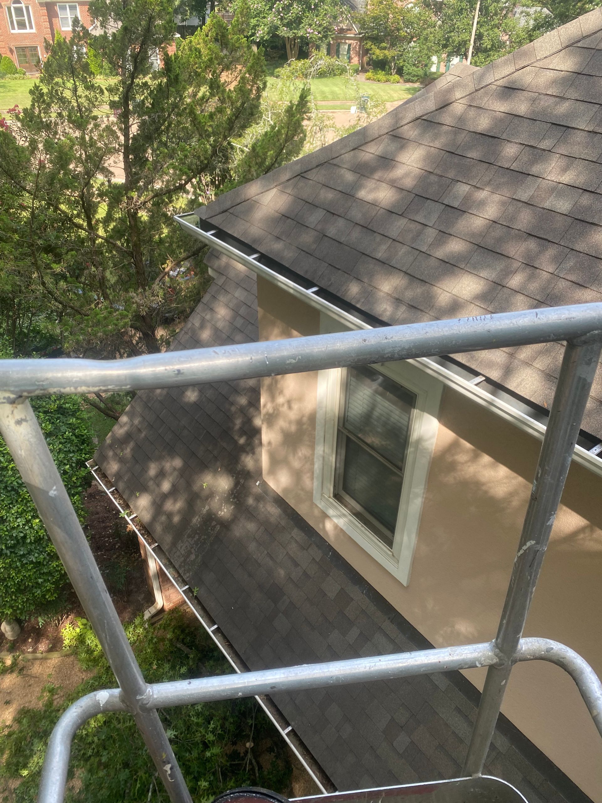 View from a ladder of a beige house with a dark roof and gutters. A window is visible, surrounded by white trim.