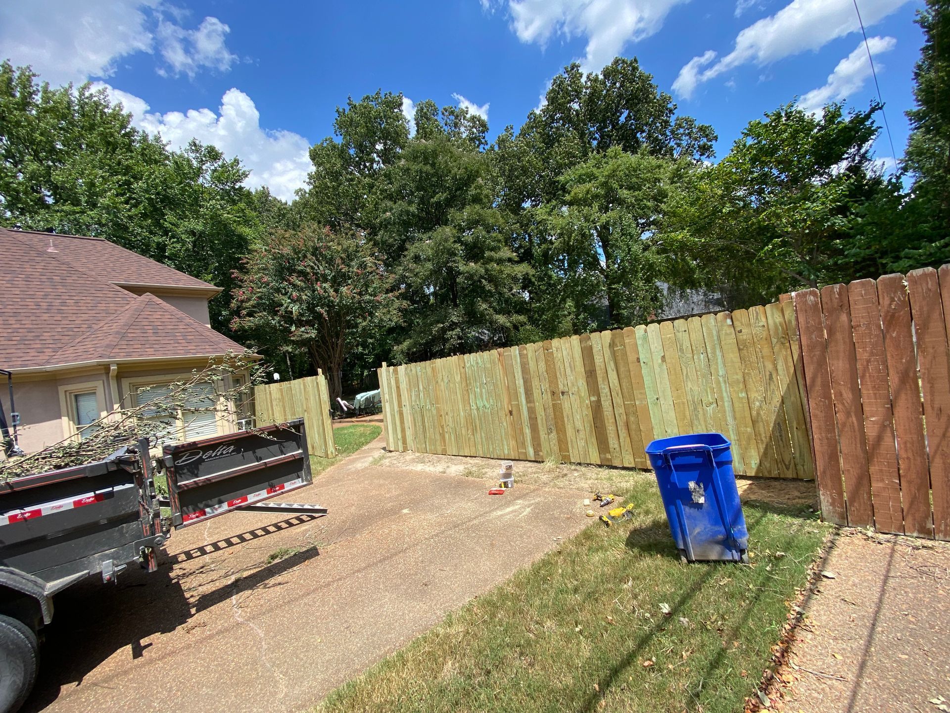 New wooden fence alongside a driveway, with a blue trash bin nearby.  A truck is parked on the left, with green grass and trees in the background.