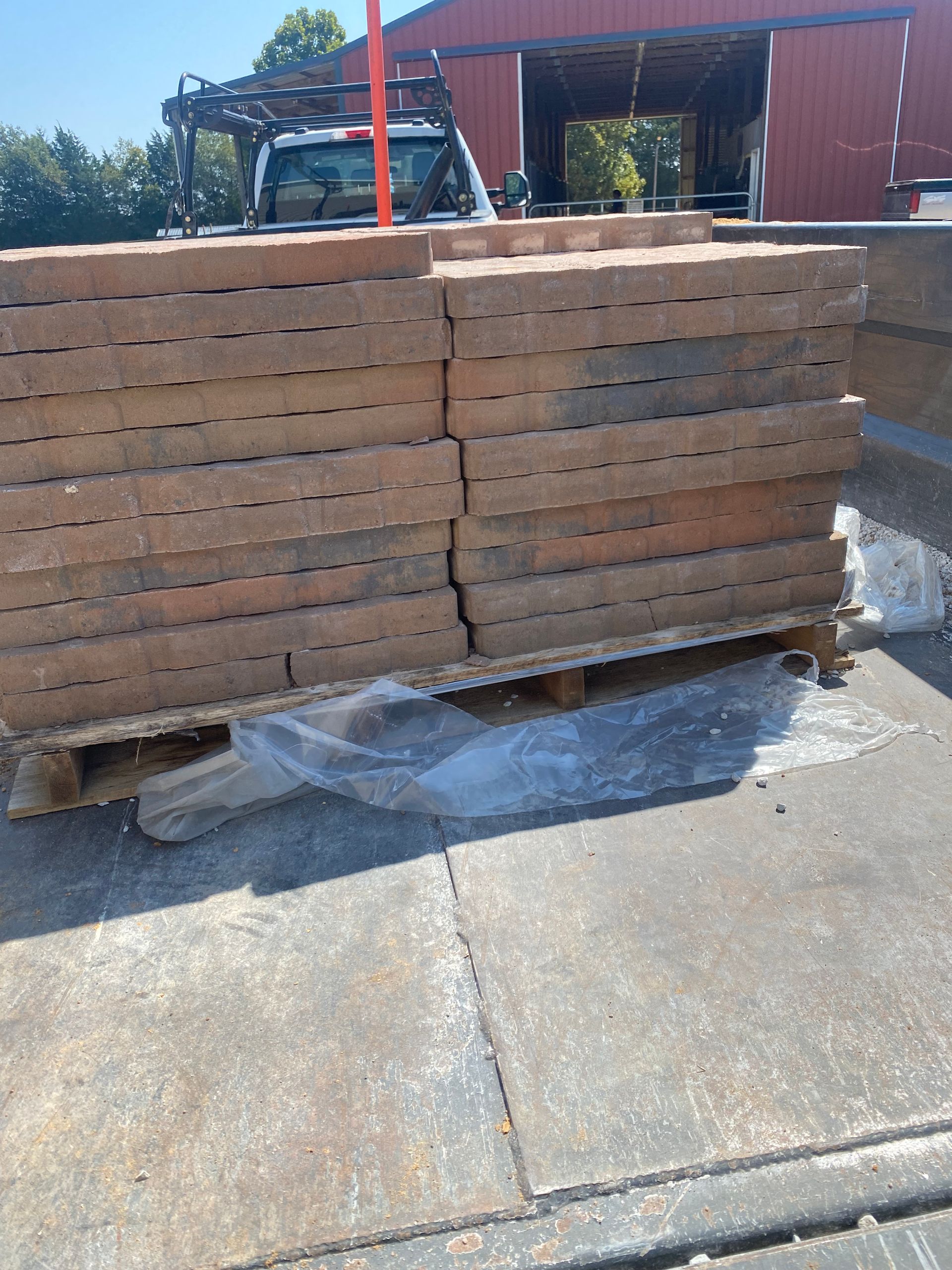 A pallet of rectangular, reddish-brown bricks wrapped in plastic, parked outside a red building with a white truck in the background.