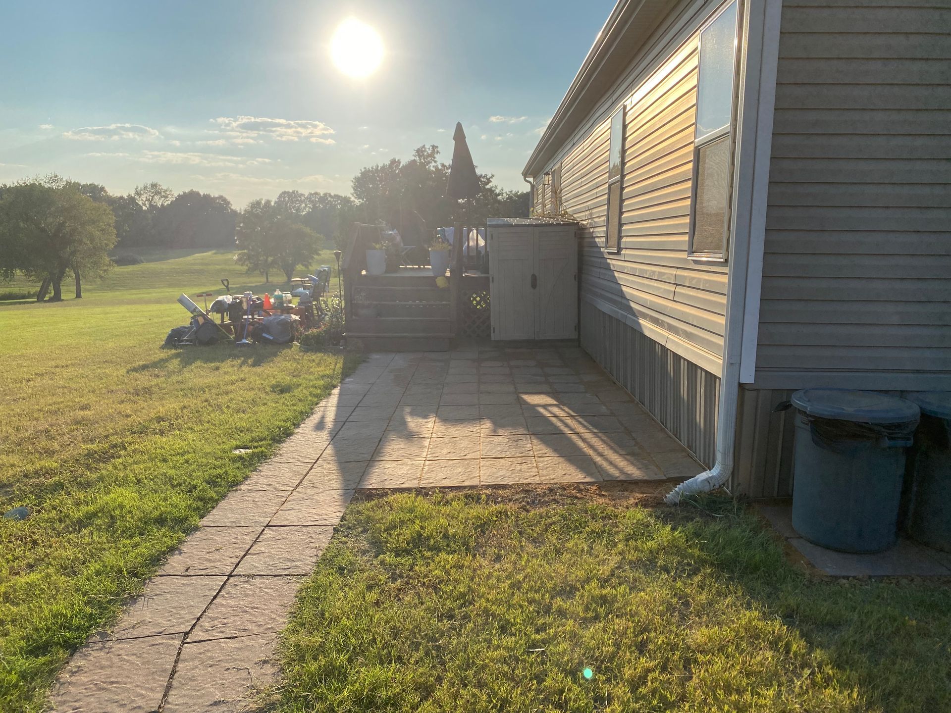 A sunny backyard scene with a house on the right, a concrete patio, and a grassy lawn leading to a treeline.