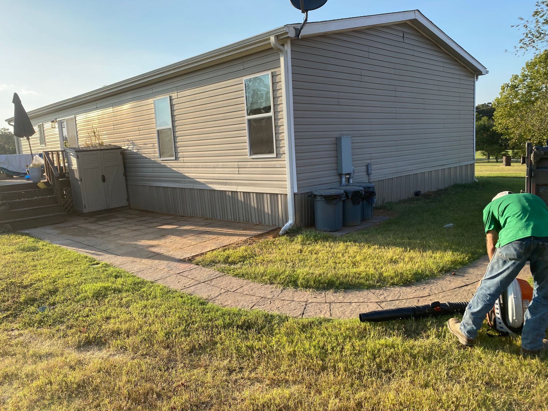 A person using a leaf blower on a grassy lawn next to a tan mobile home on a sunny day.
