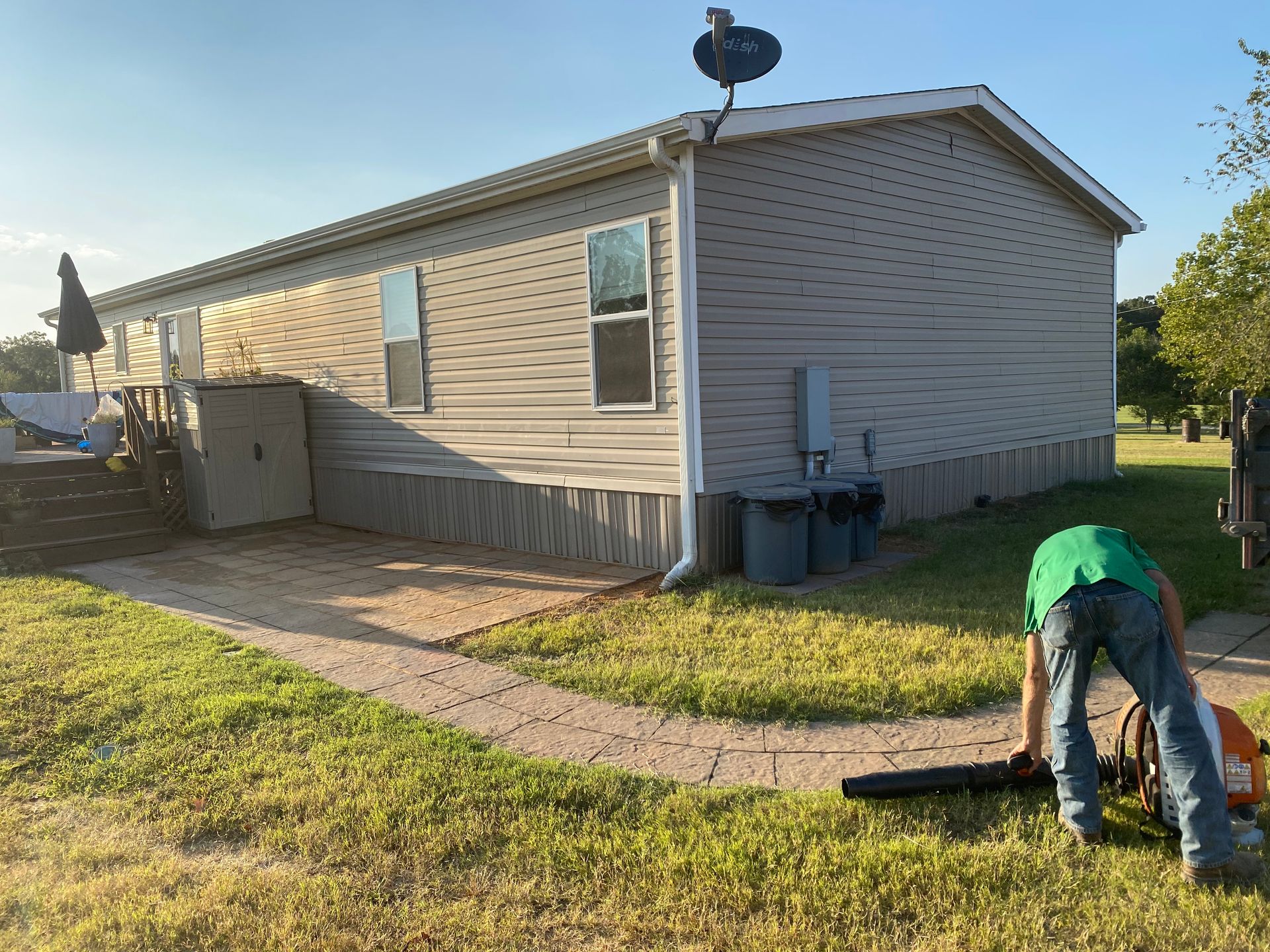 A person in a green shirt uses a leaf blower in a yard next to a light gray mobile home with a satellite dish.