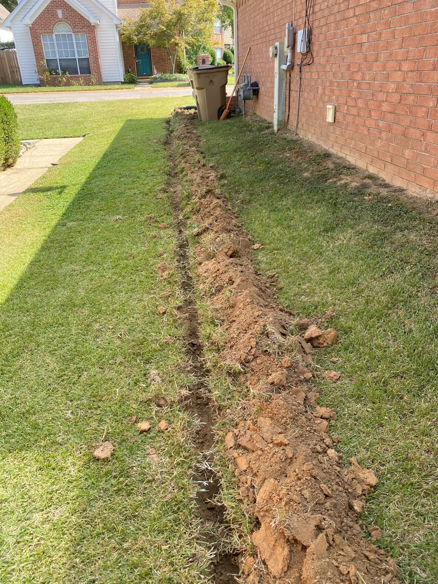 Trench dug in a grassy yard next to a brick building. Brown soil is piled along the edge of the trench.