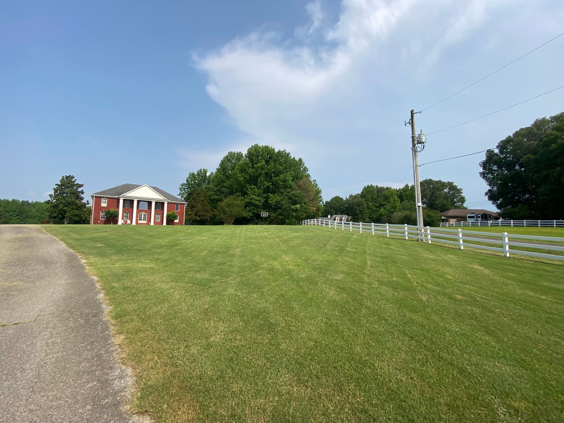 A large red brick house with white columns stands in a field, a paved driveway to the left. White fences run parallel to the house.