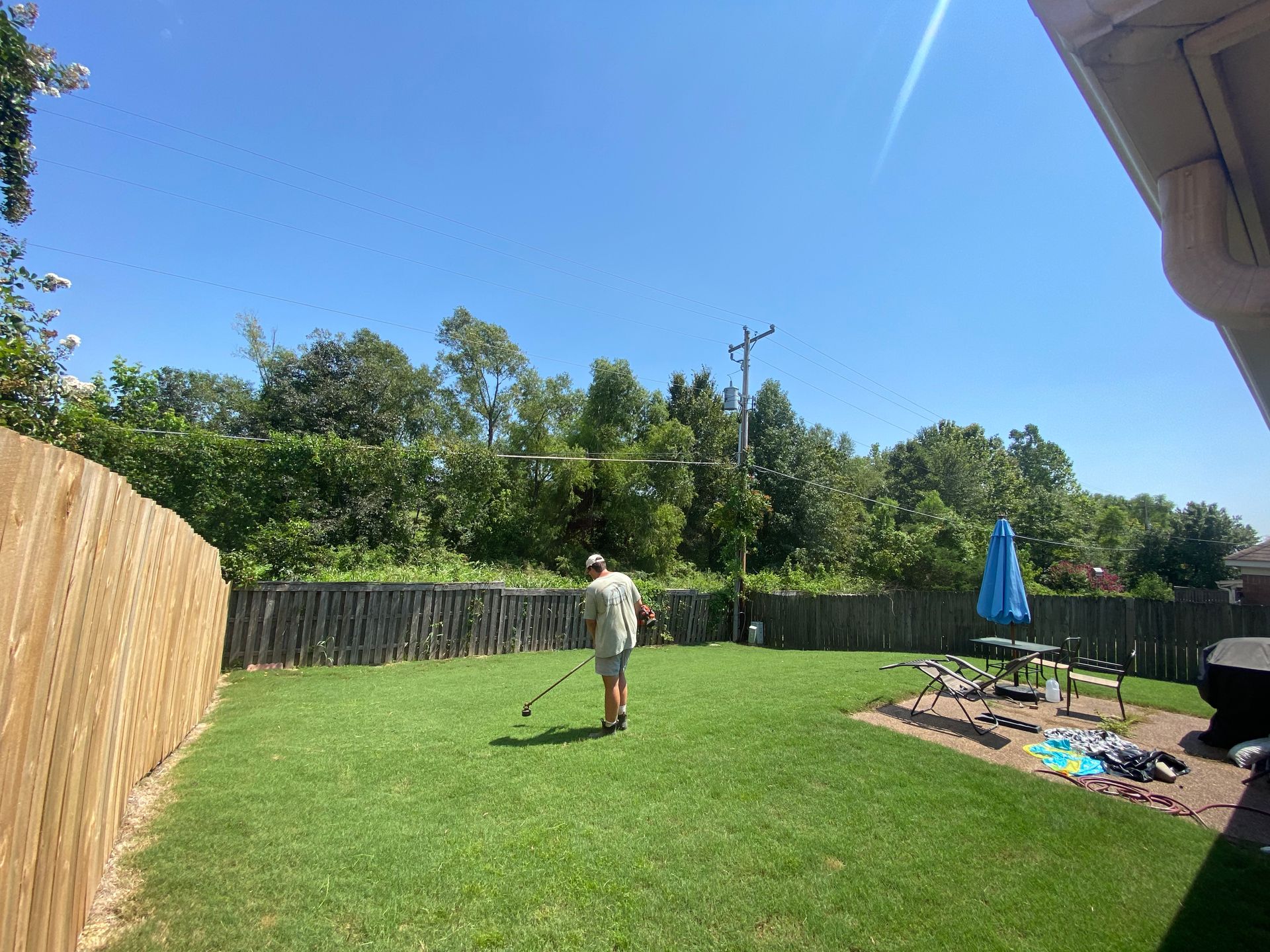 Man in backyard, holding a golf club, with green grass, blue sky, and a wooden fence.