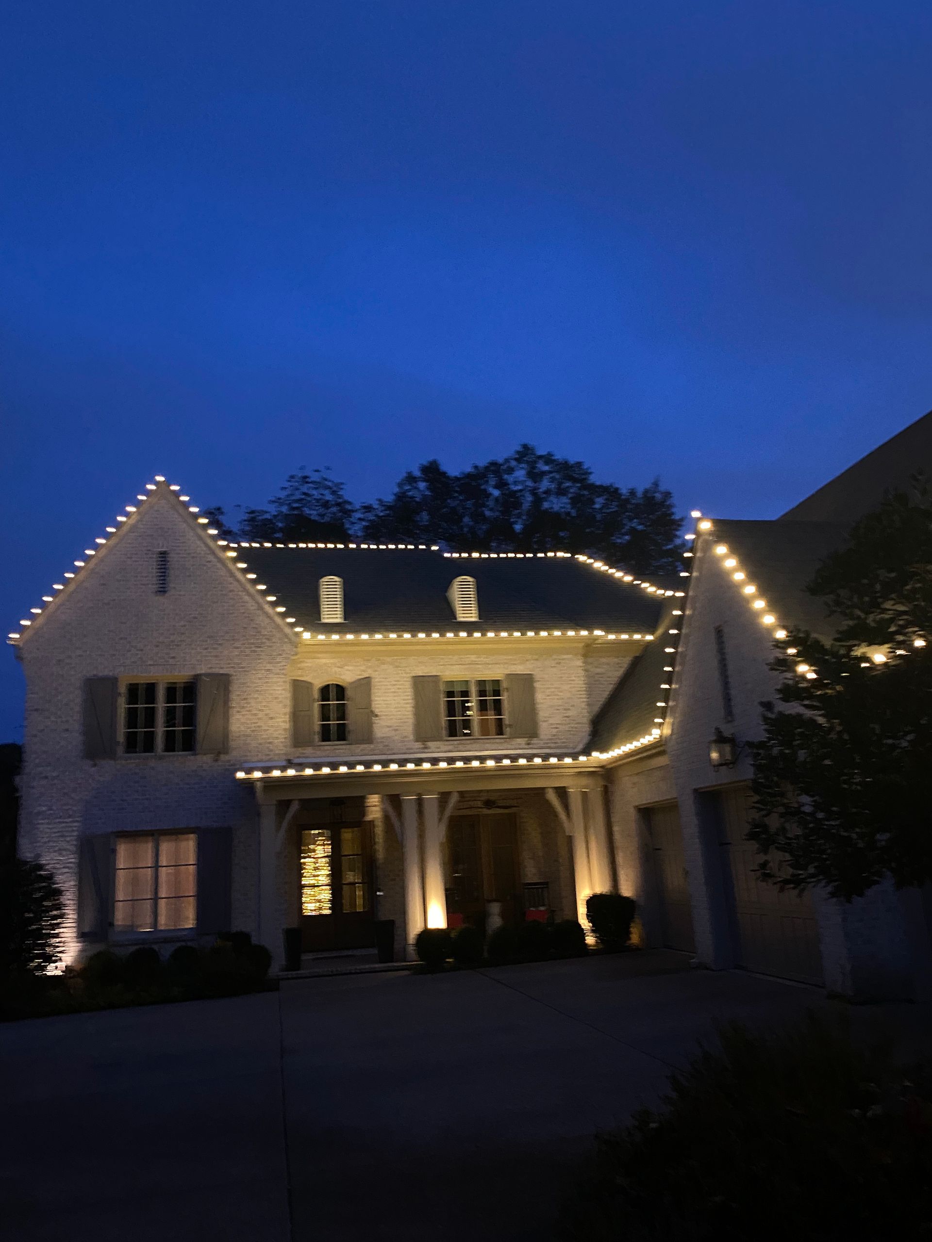 A two-story house at dusk, decorated with white lights along the roofline. The front door is illuminated, and a Christmas tree can be seen inside.