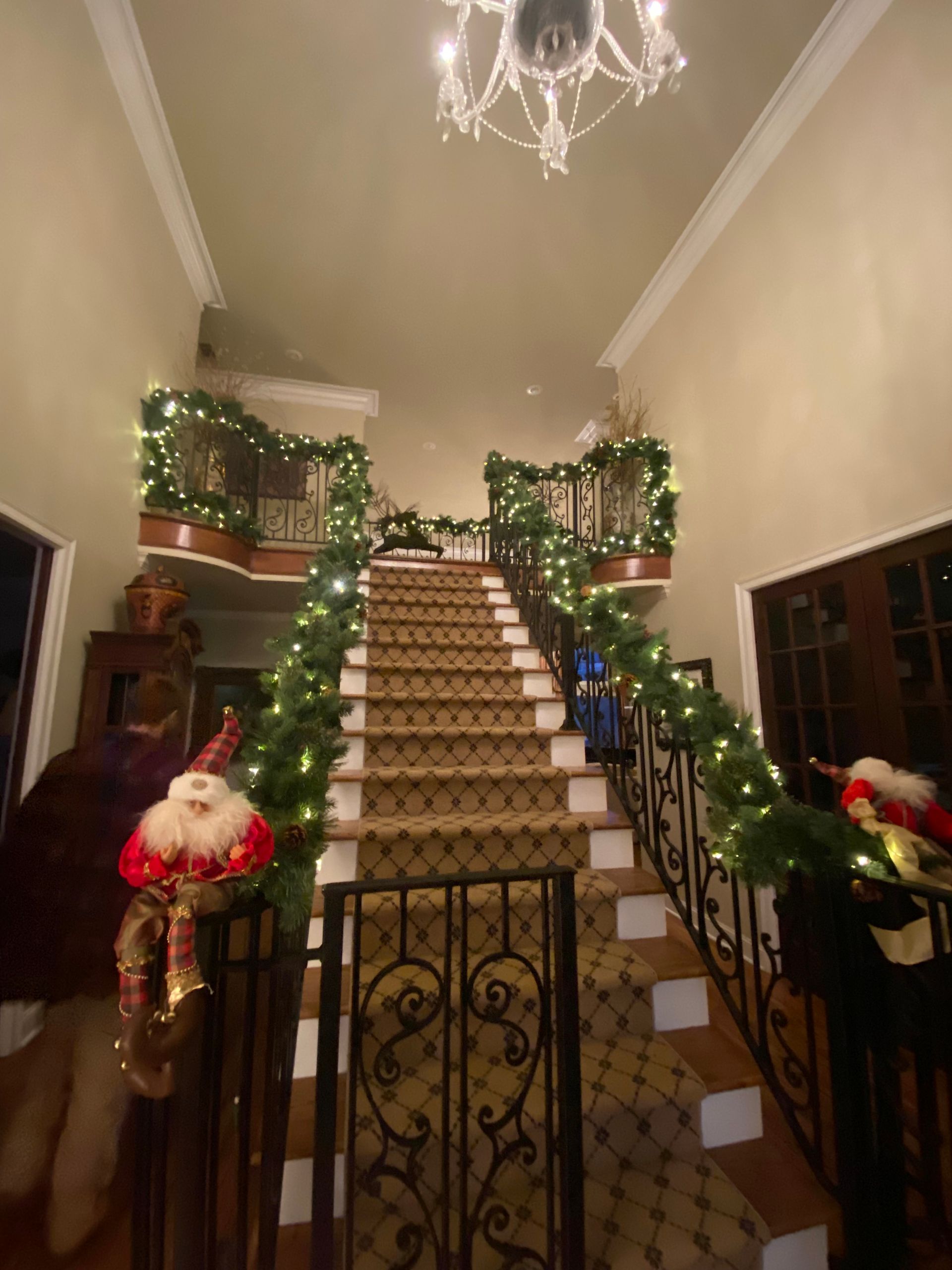 A grand staircase decorated for Christmas with garland, lights, and Santa figures. The setting is an entryway with a chandelier.