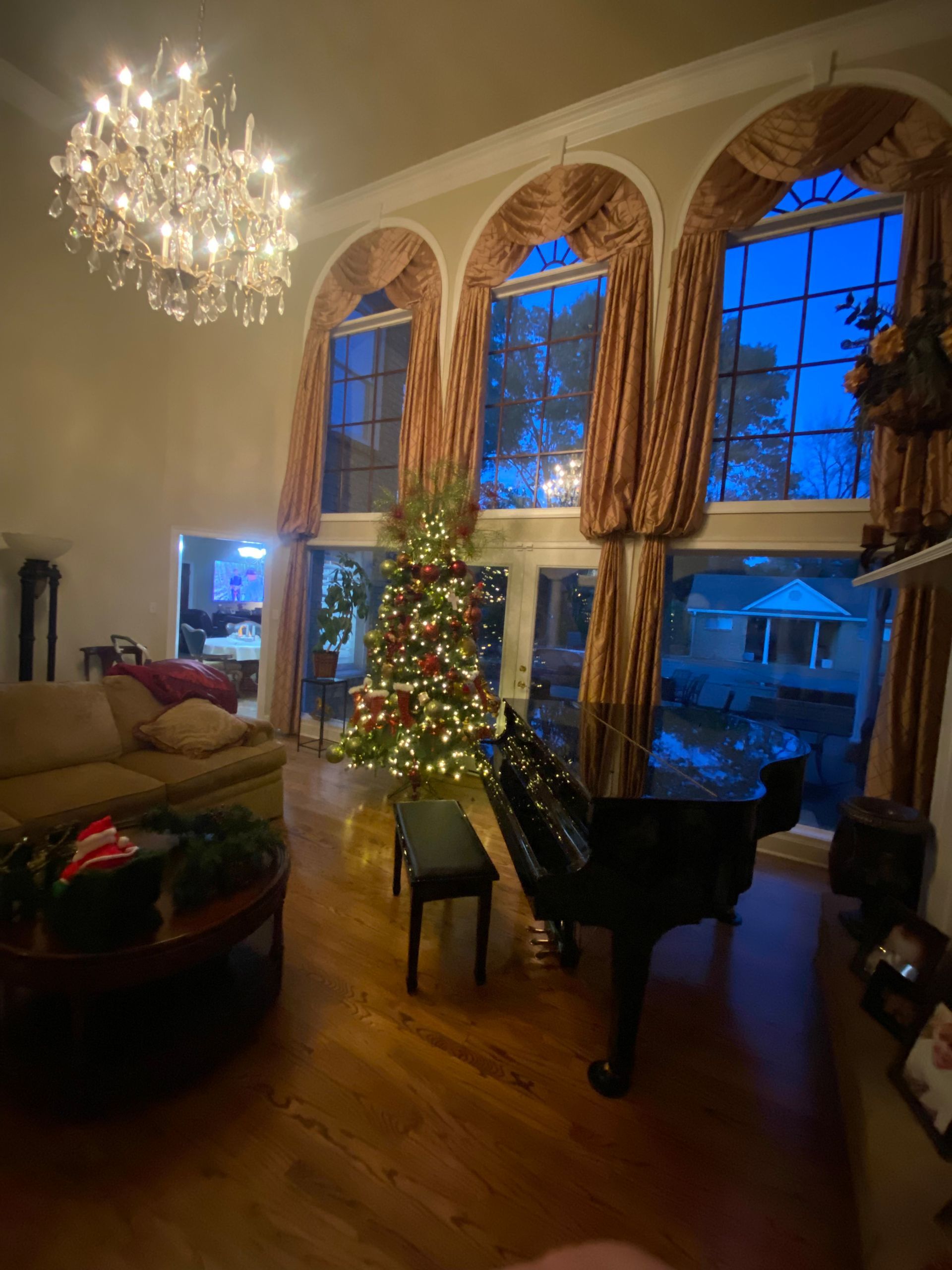 Living room decorated for Christmas with a grand piano, Christmas tree, and large arched windows overlooking a blue-lit outdoor view.