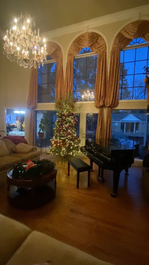 A brightly lit living room with a Christmas tree and a grand piano. Large windows with draping curtains frame the outside view.