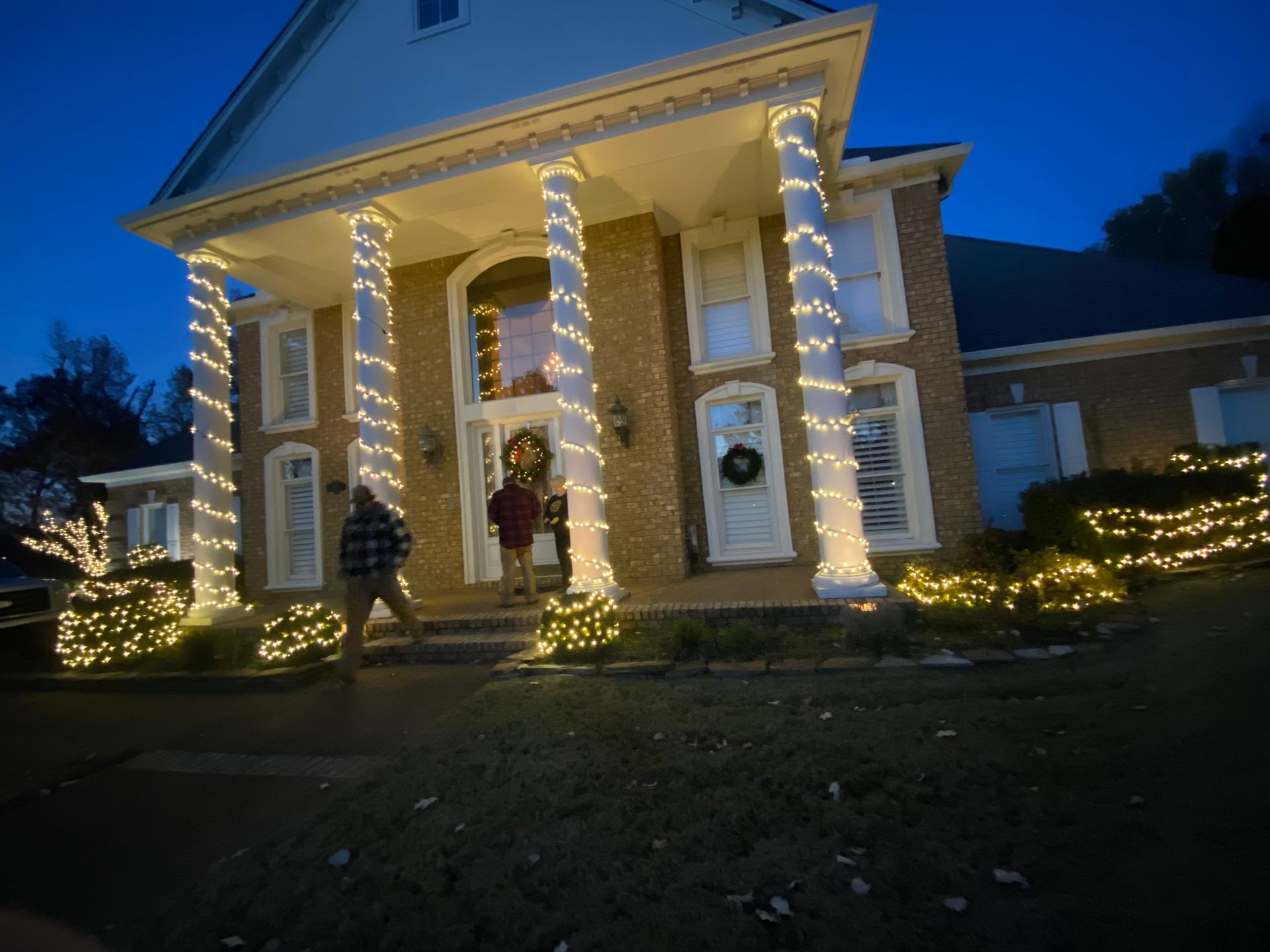 House decorated with Christmas lights, featuring people walking towards the front door. Blue sky backdrop.