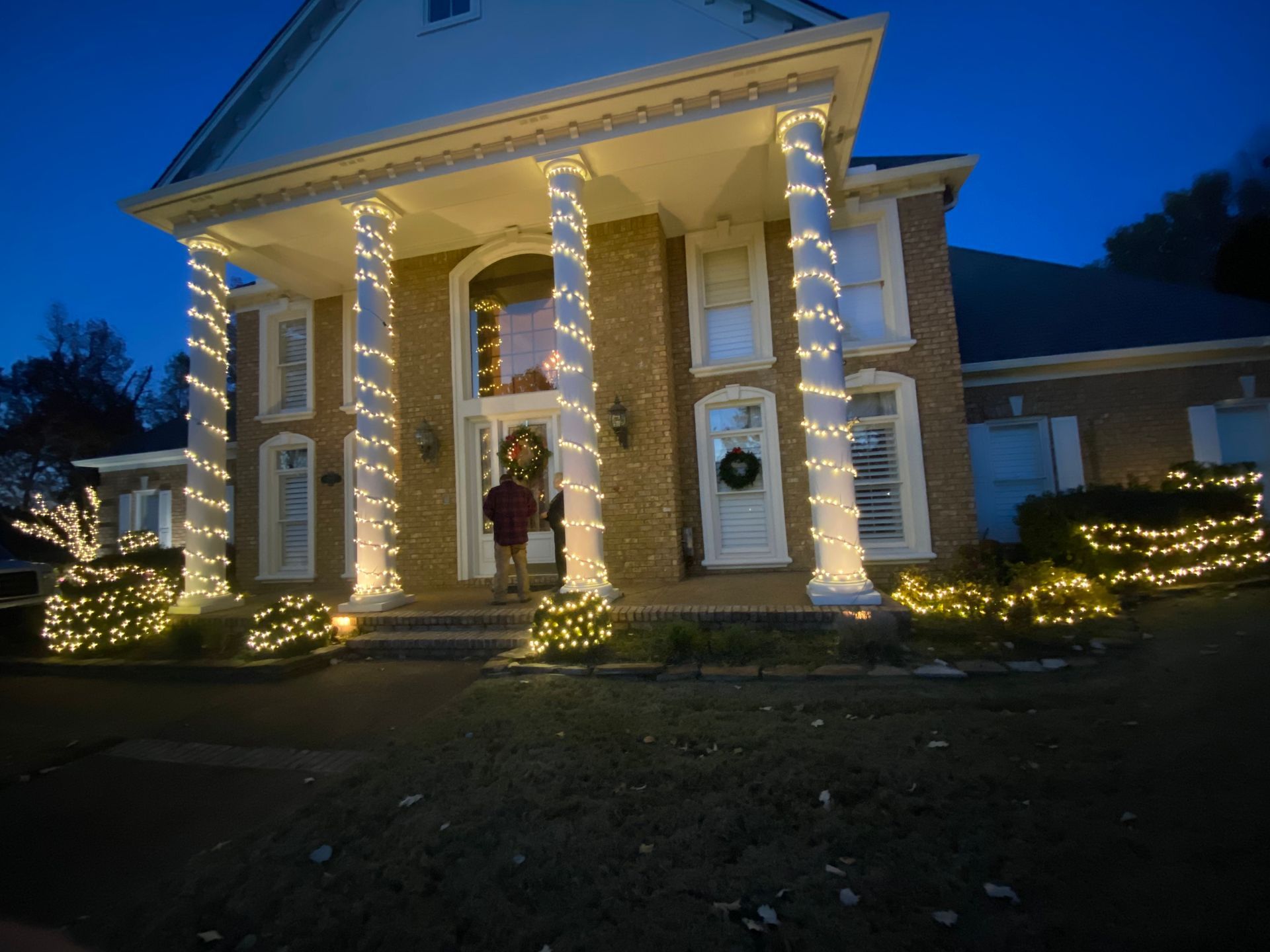 A two-story brick house decorated with Christmas lights. Columns and bushes are wrapped in white lights; wreaths adorn the front door.
