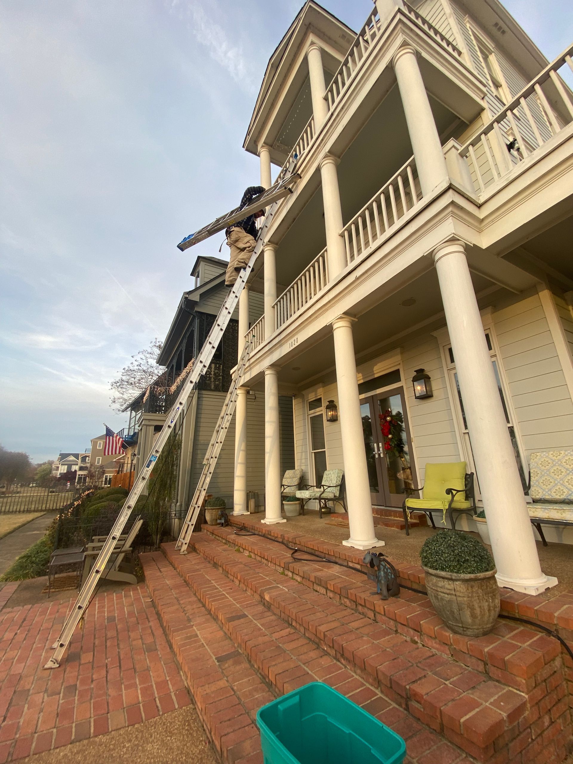 A person on a tall ladder works on the roof of a two-story white house with red brick steps. A blue bin sits below.