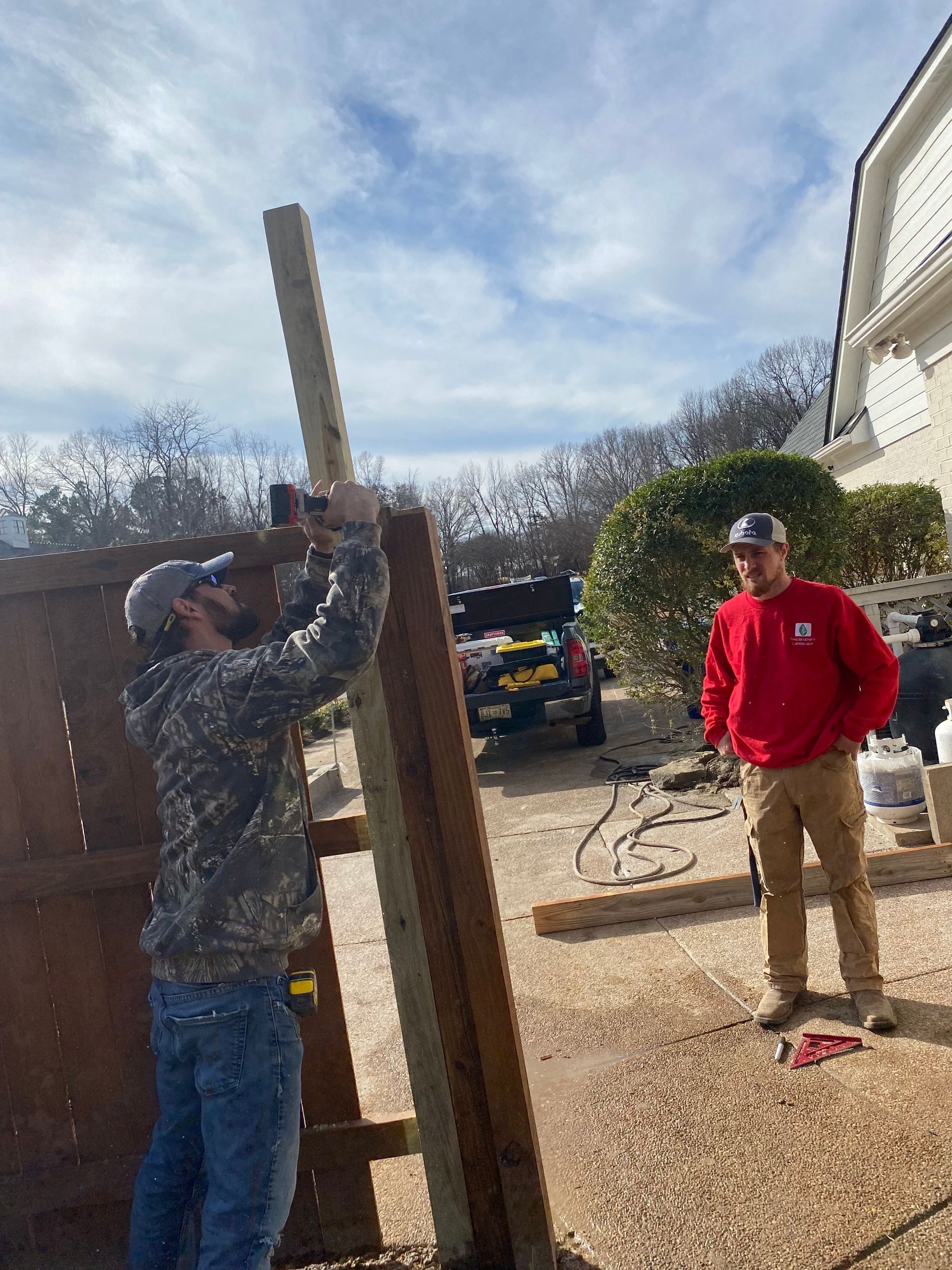 Two men building a fence outdoors. One uses a hammer on a wooden post while the other observes, wearing a red shirt and tan pants.
