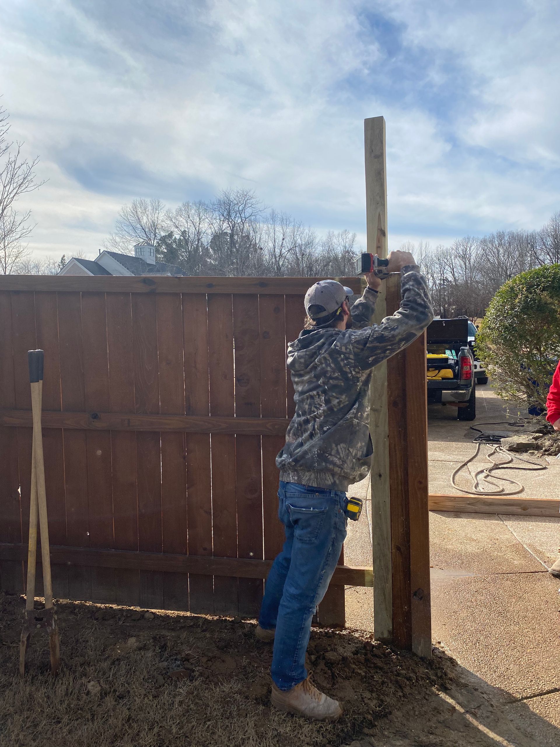 Man in camouflage jacket installing a wooden fence post. He’s using a hammer against the post, near a completed brown fence.