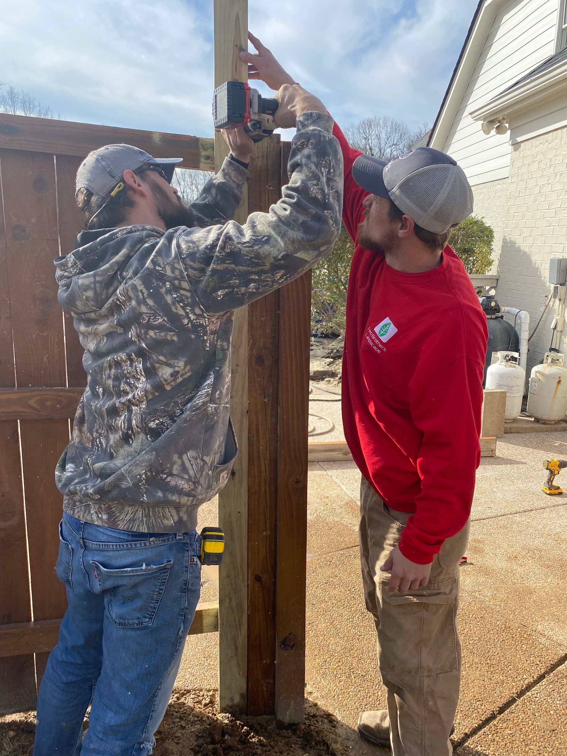 Two men in work clothes installing a wooden fence post outdoors. One uses a tool while the other observes and assists.