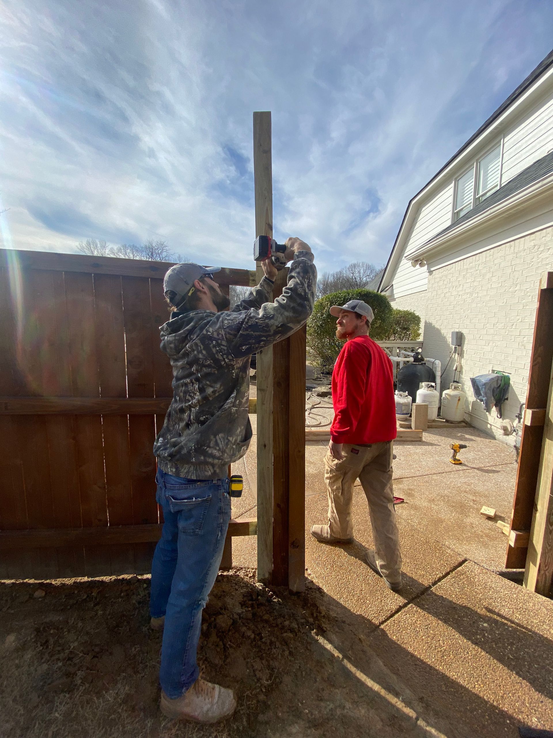 Two men building a fence outdoors. One uses a tool on a post, the other watches. Blue sky overhead.