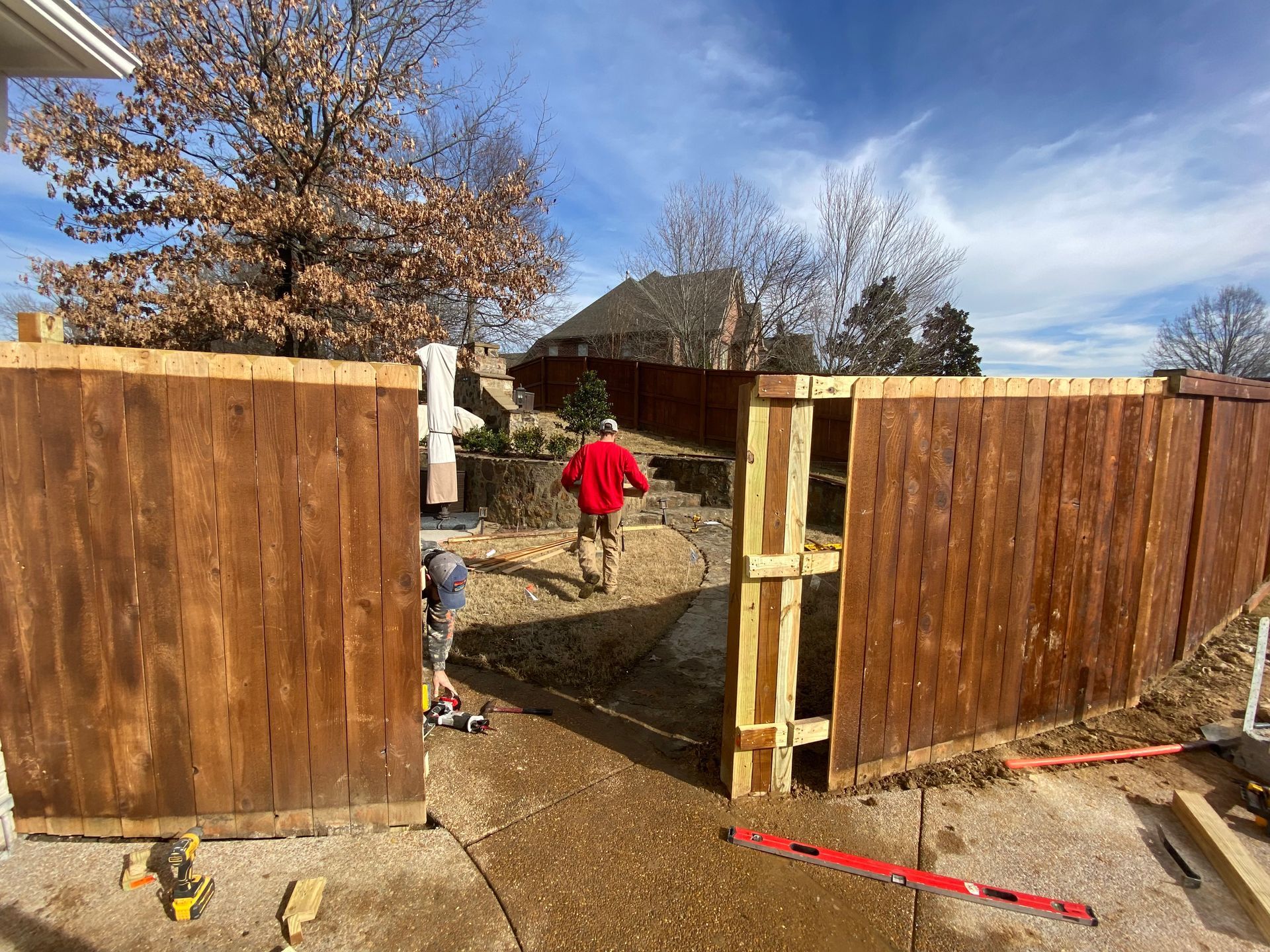 Workers constructing a wooden fence with a gate. A person in a red jacket walks through the opening towards a house under a blue sky.