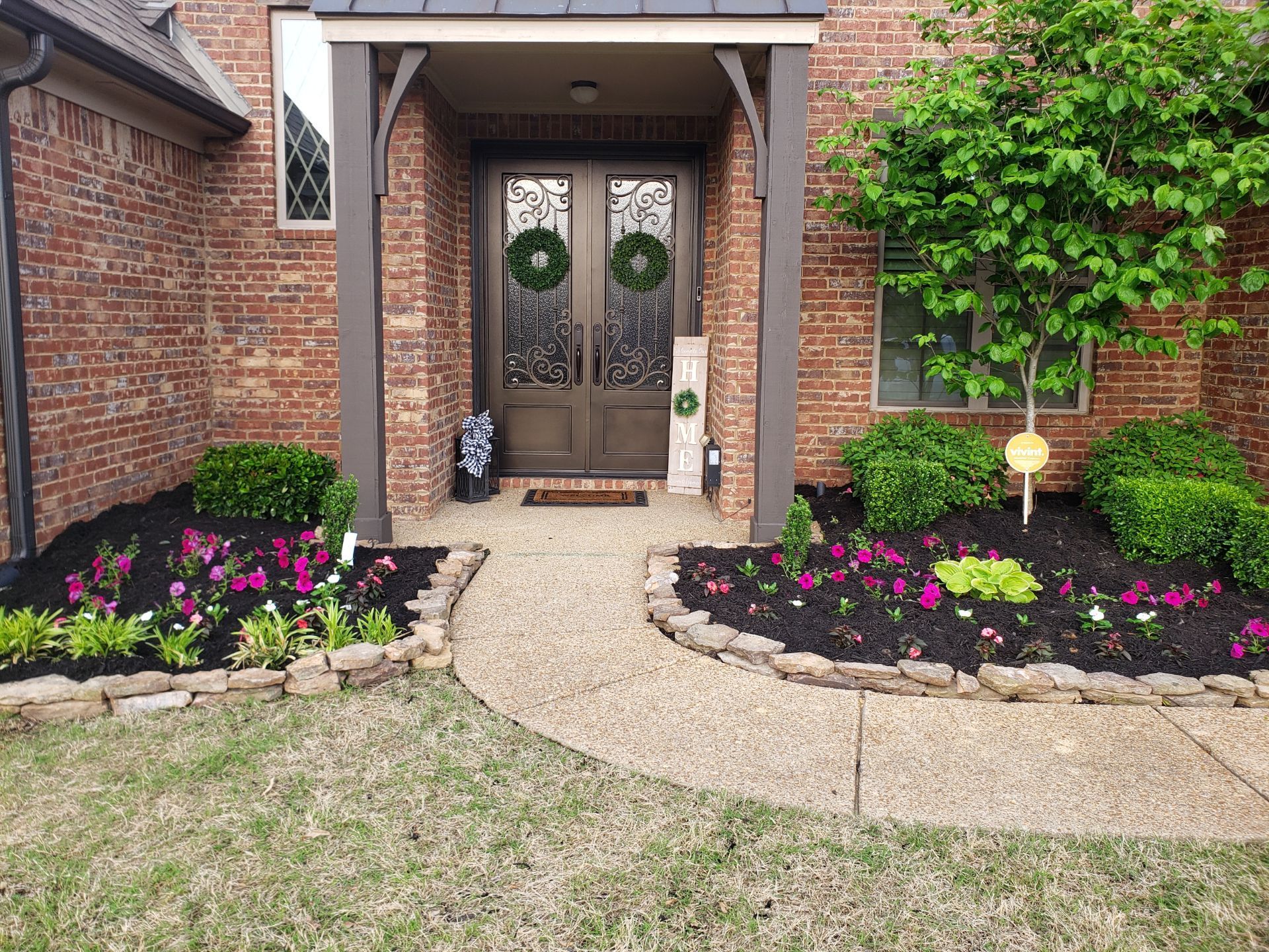 Brown brick home with a walkway leading to double doors. The doors are decorated with wreaths and flanked by landscaped flowerbeds with pink and white flowers.