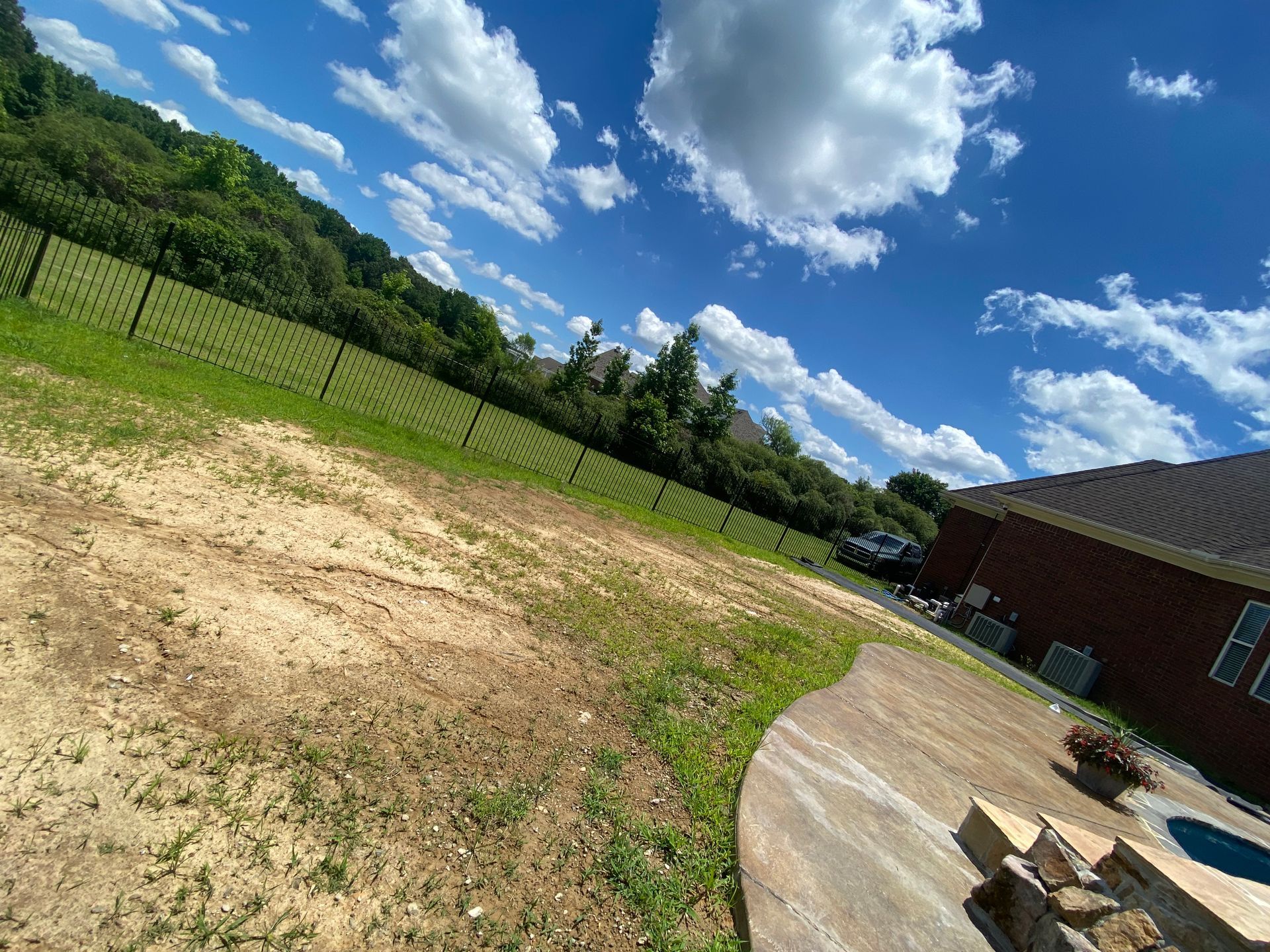 Backyard with bare earth and patchy grass, a fence, and a house under a blue sky with fluffy white clouds.