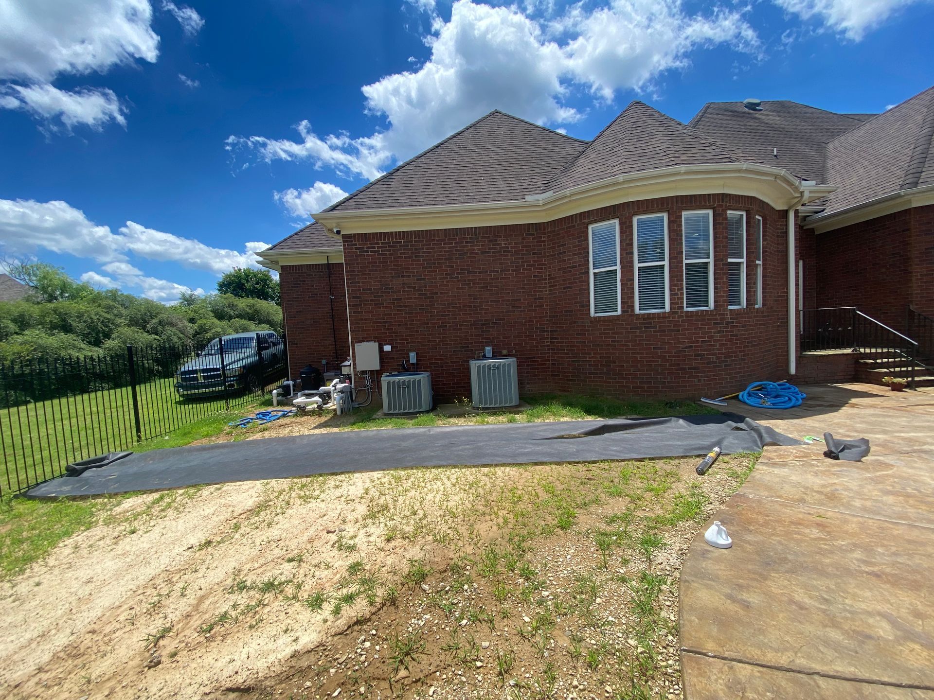 A brick house with brown roof, two AC units, and a paved driveway on a sunny day. A truck sits in the background.