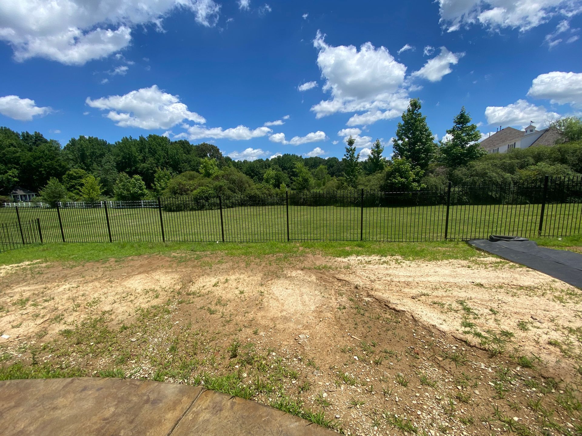 A backyard with a sandy area, grass, and a black fence against a backdrop of trees and a blue sky with clouds.