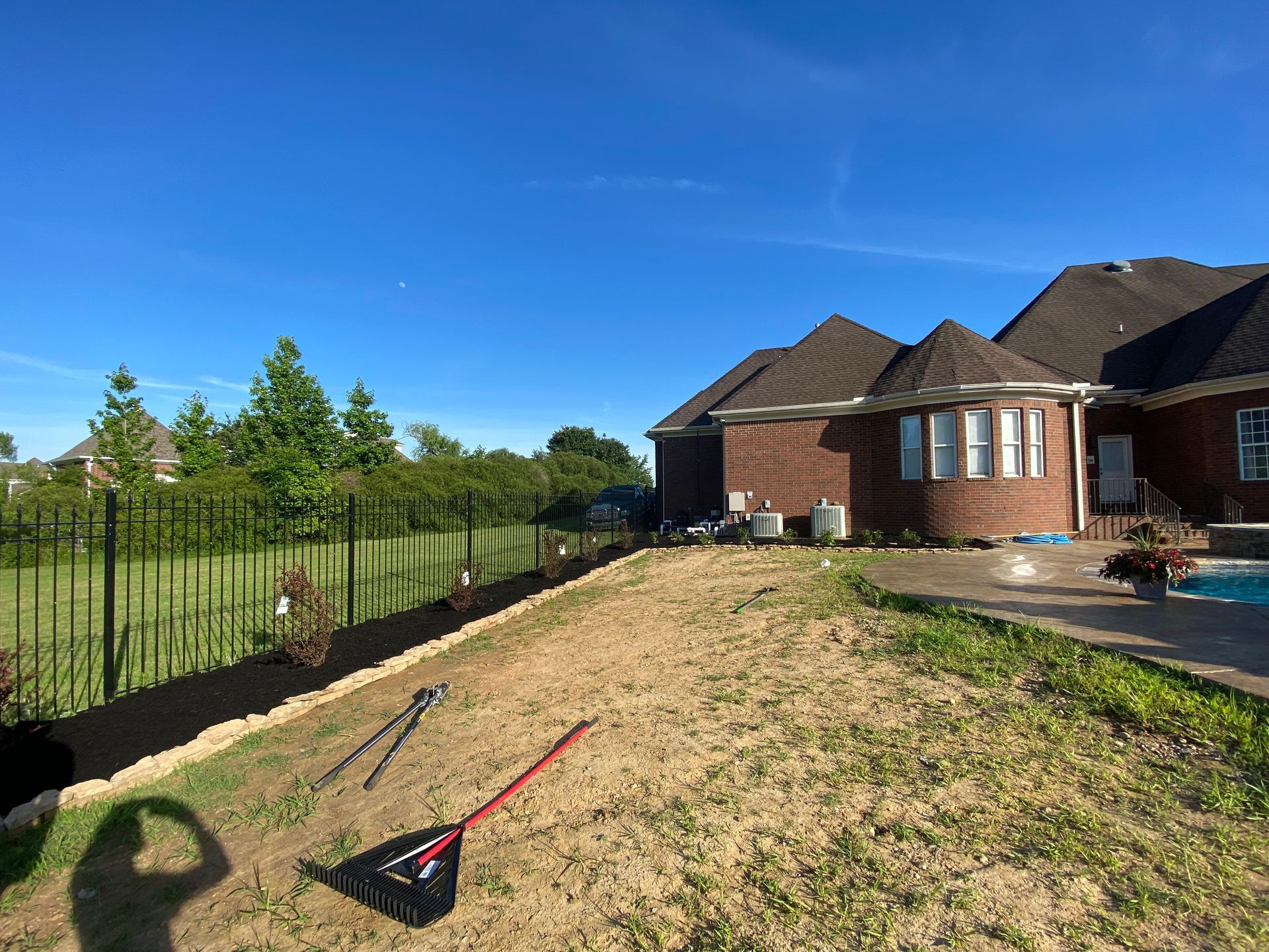 Backyard with a house and fence under a blue sky. A bare patch of dirt is in front of the house, and some tools are resting on the ground.