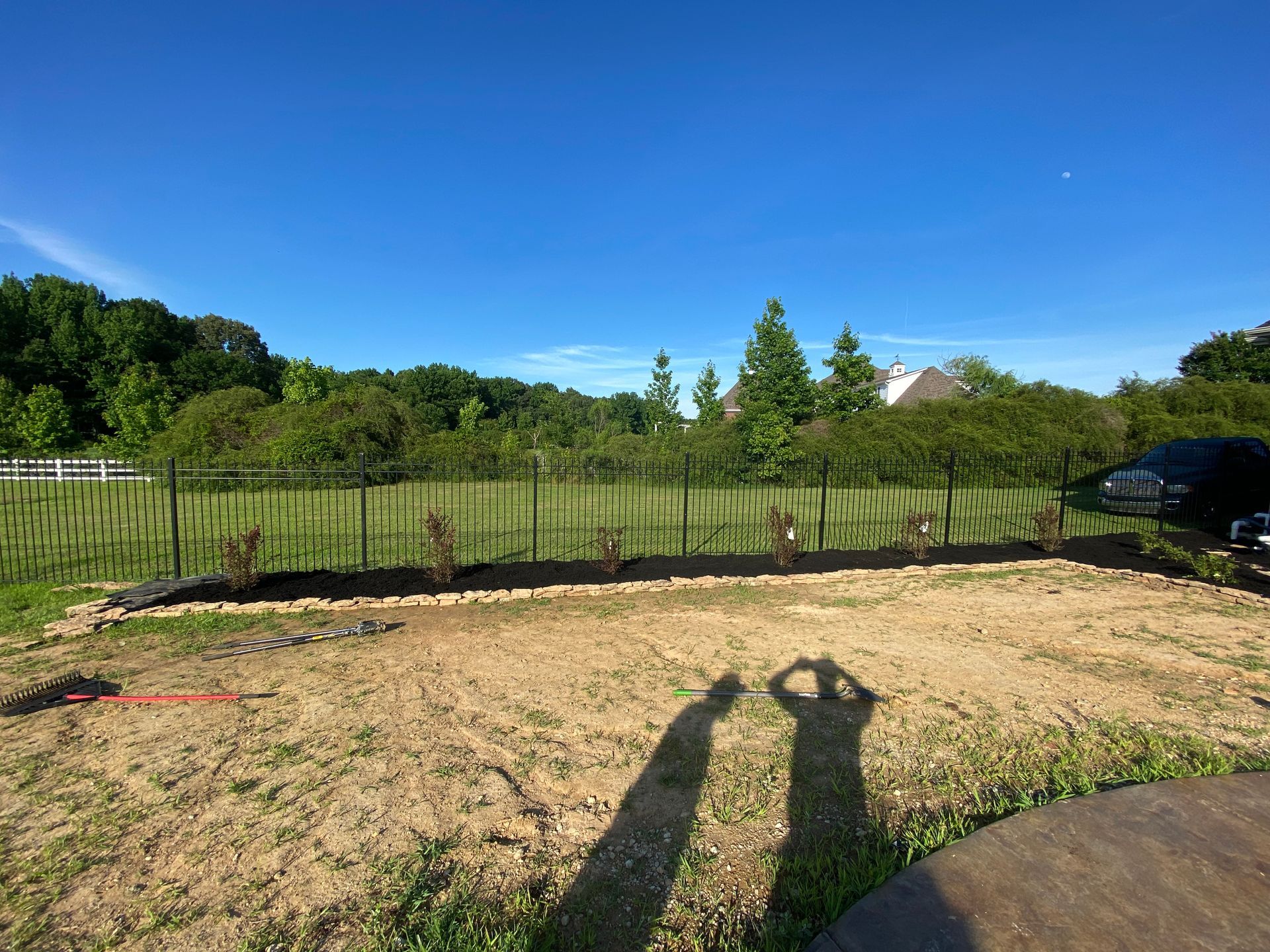 Sunny outdoor scene with a freshly tilled garden bed in front of a black fence and treeline under a bright blue sky.