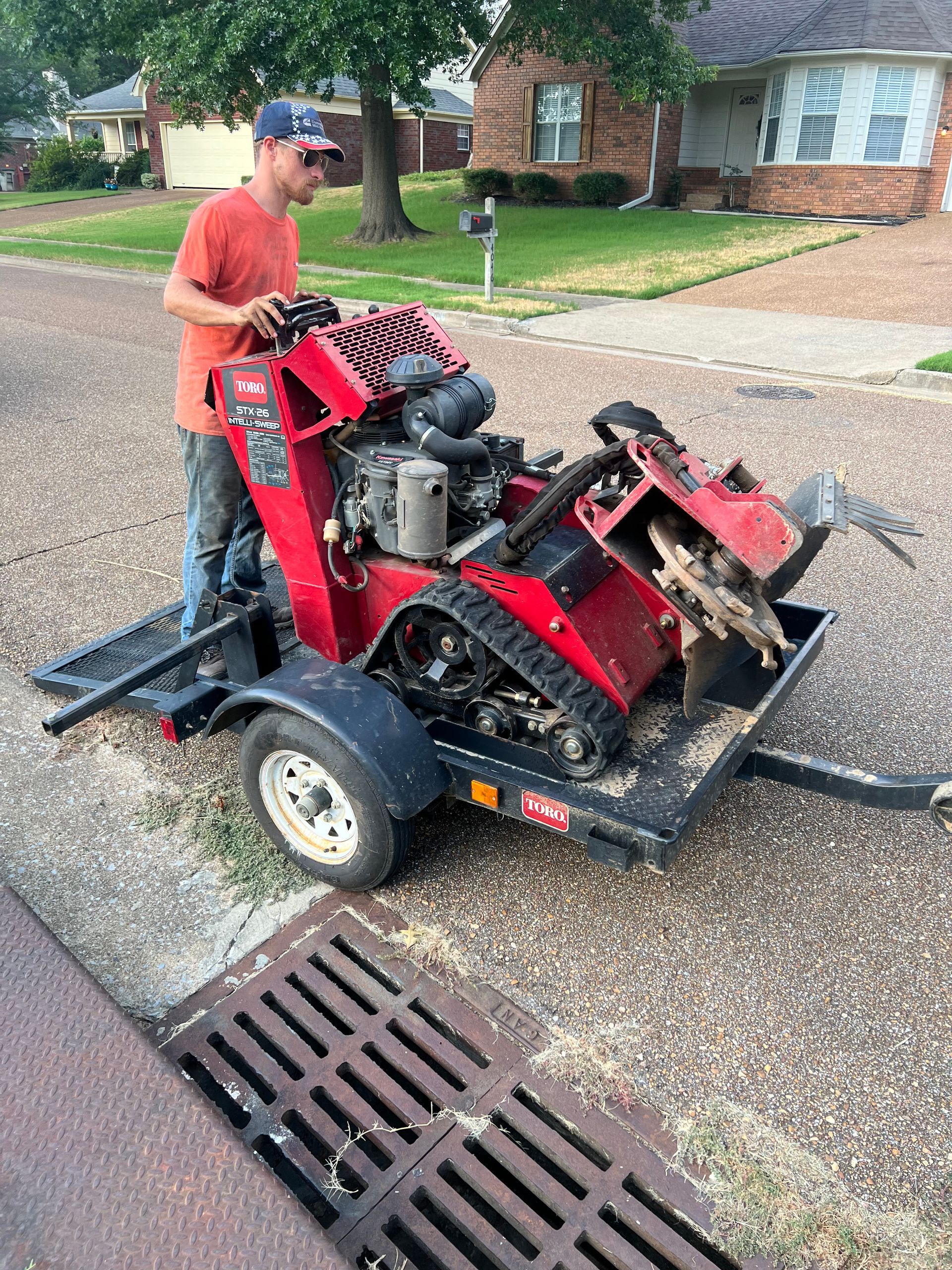 Man operating a stump grinder on a trailer by a curb. The machine is red and black, and wood chips are scattered.