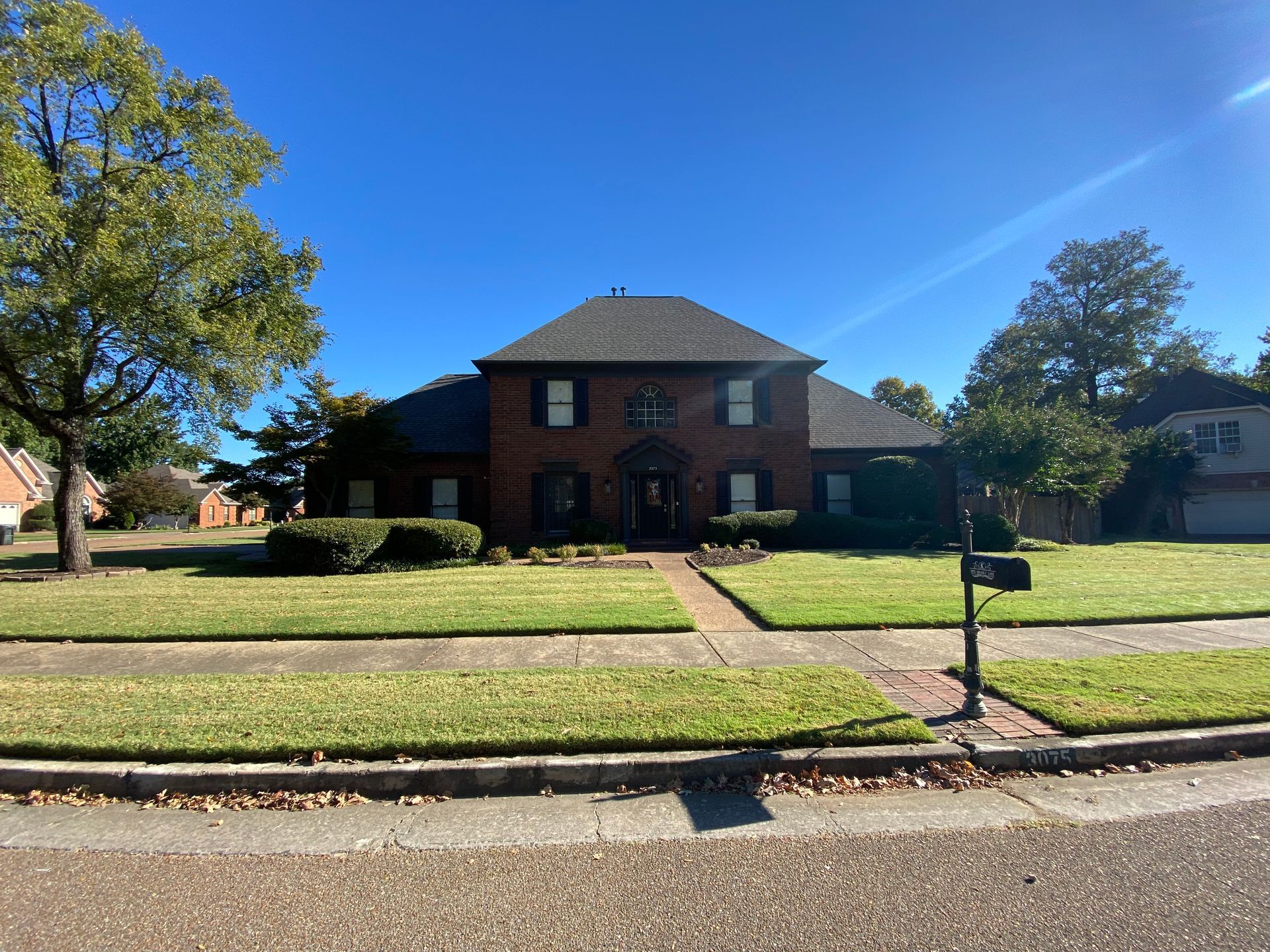 A brick house with a dark roof and green lawn under a clear blue sky. A mailbox and trees are in the yard.