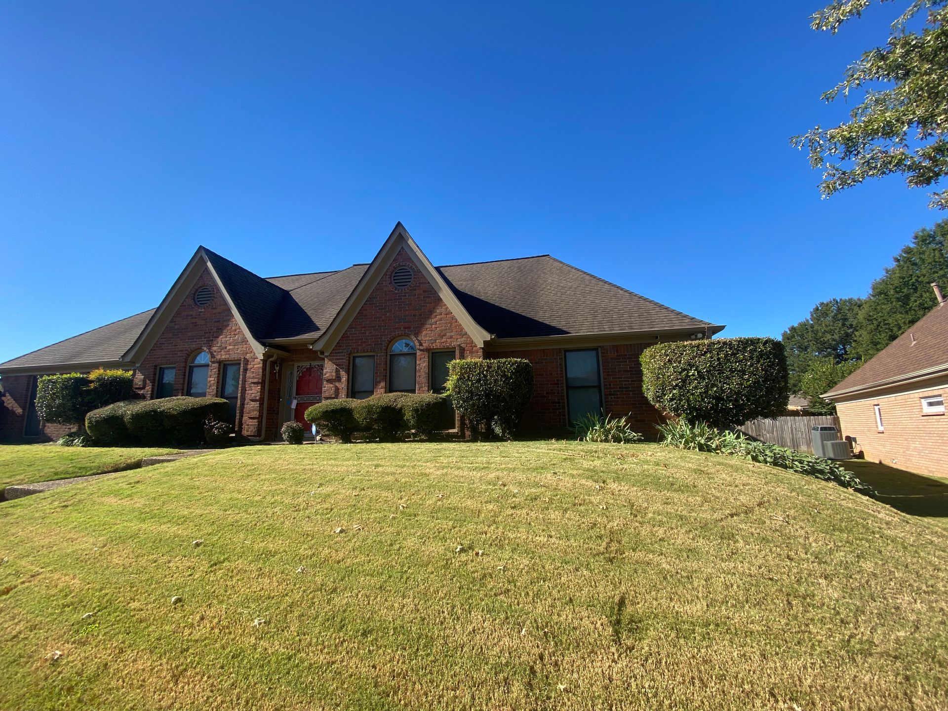 Brick house with dark roof, trimmed bushes, and a grassy lawn under a clear blue sky.