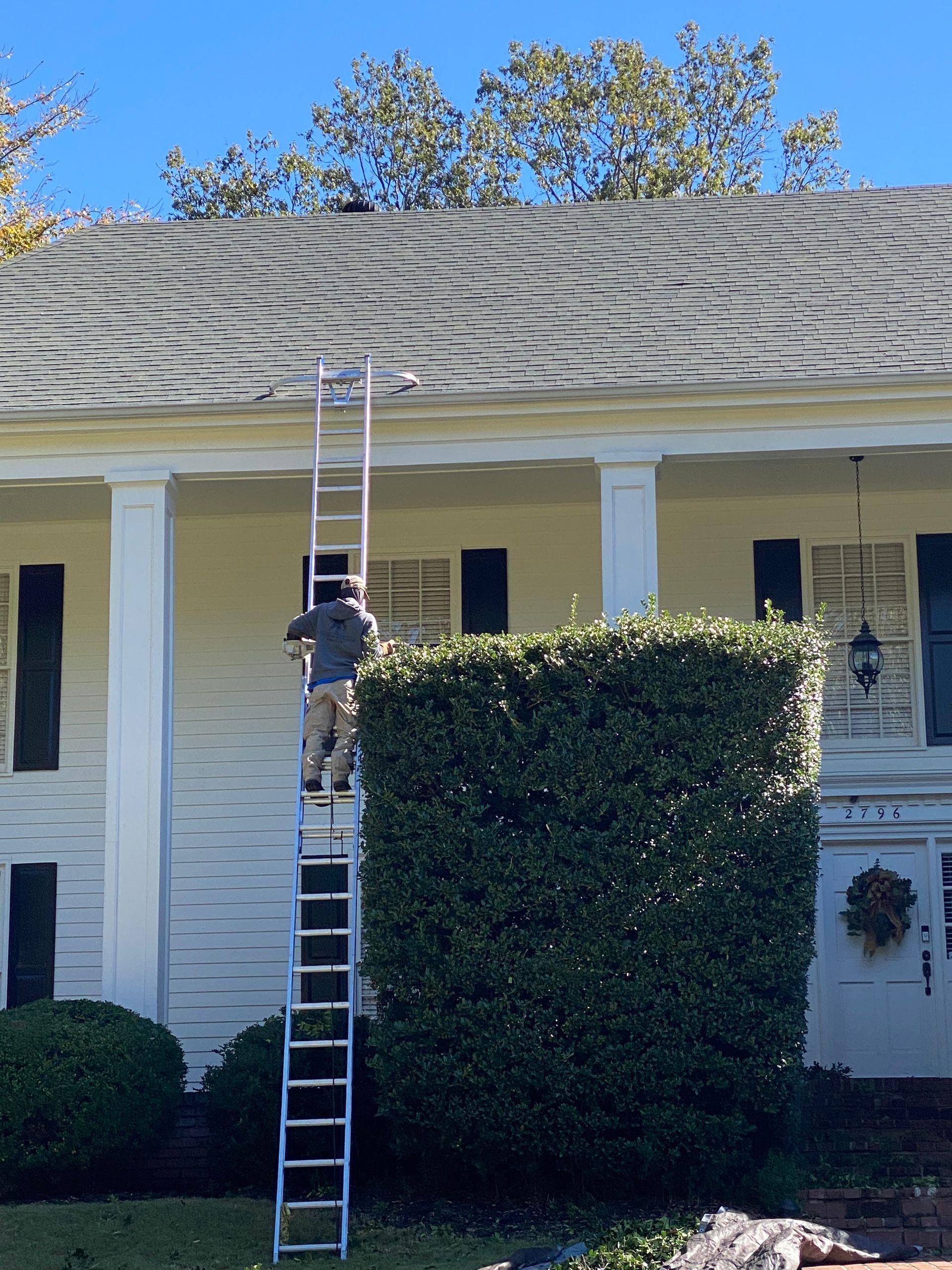 Person on a tall ladder cleaning a roof gutter on a white house with columns and a front hedge.