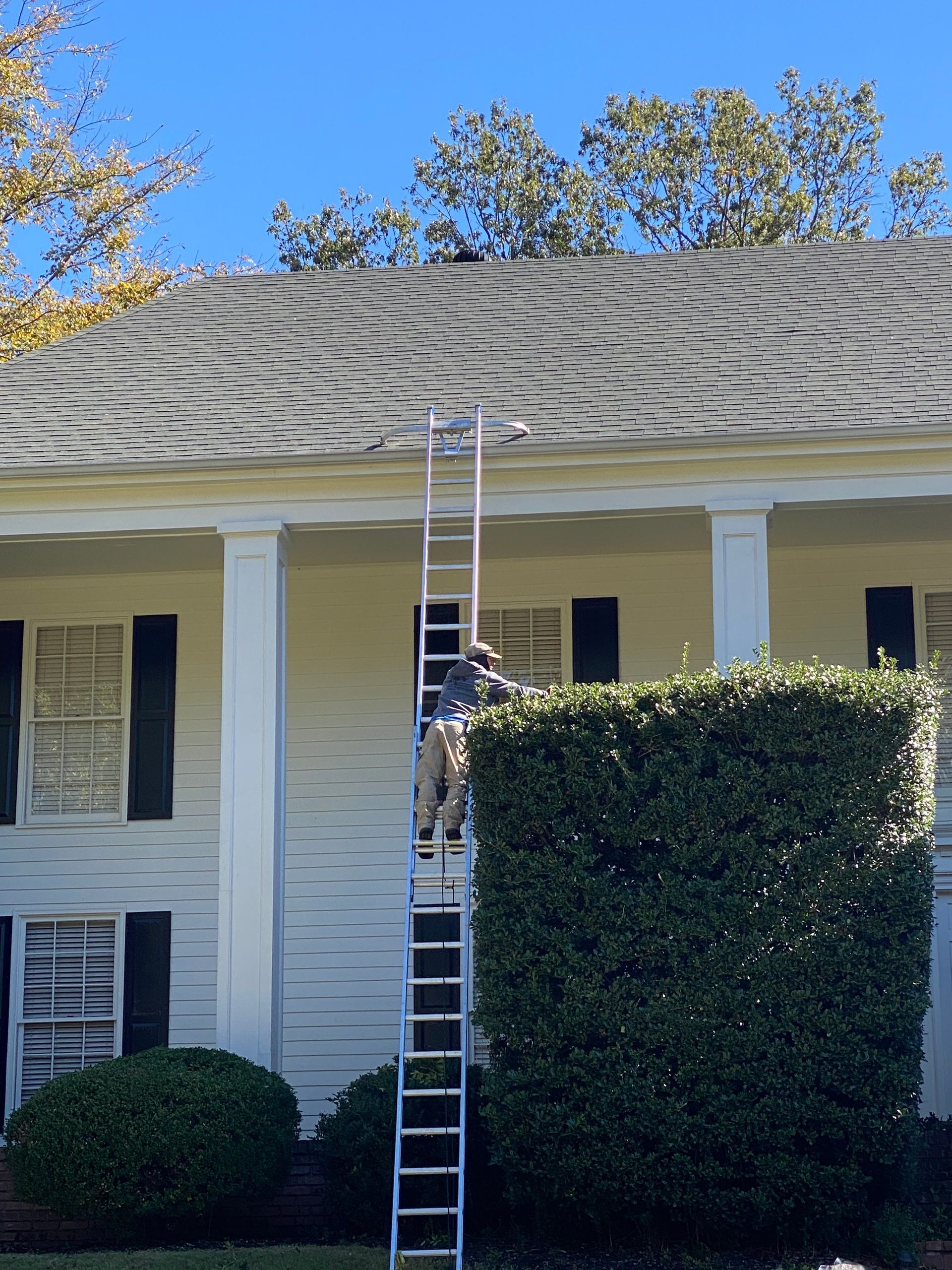 Person on a tall ladder cleaning gutters on a white house with dark shutters and a blue sky backdrop.
