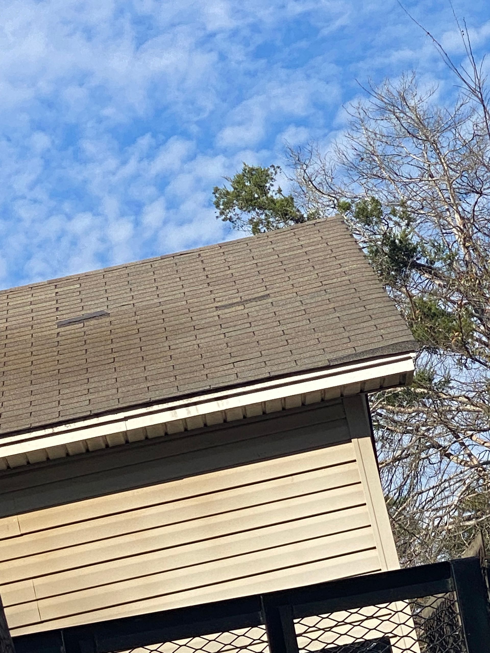 A light brown roof with dark-colored shingles sits on a tan-sided building against a blue sky dotted with clouds.