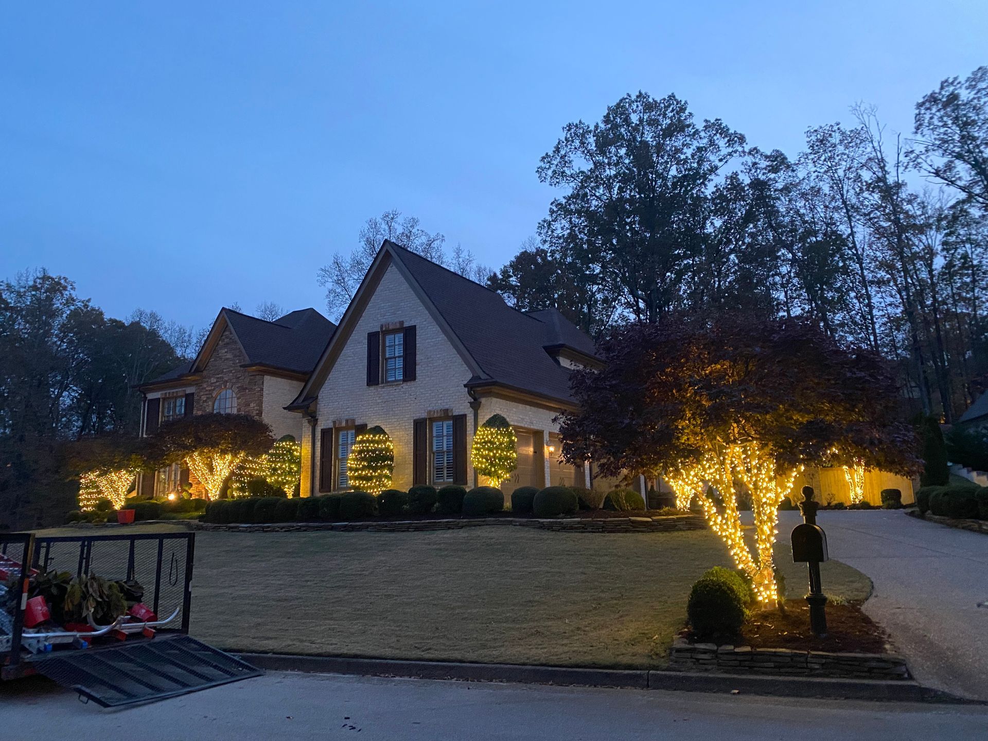 House decorated with warm white Christmas lights in the evening. Wreaths and tree branches are illuminated, with a lawn and driveway in view.