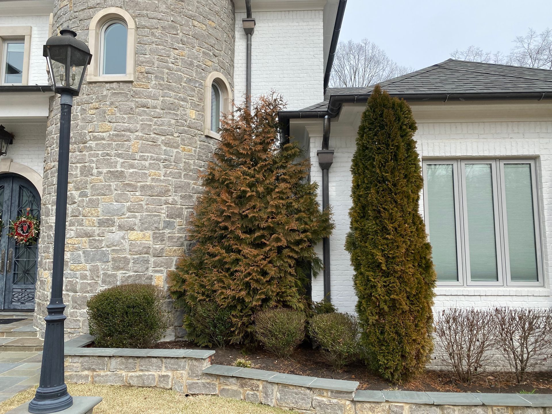 A house with a stone turret and white brick siding. Two evergreen trees are near the right side, with a lamppost on the left.