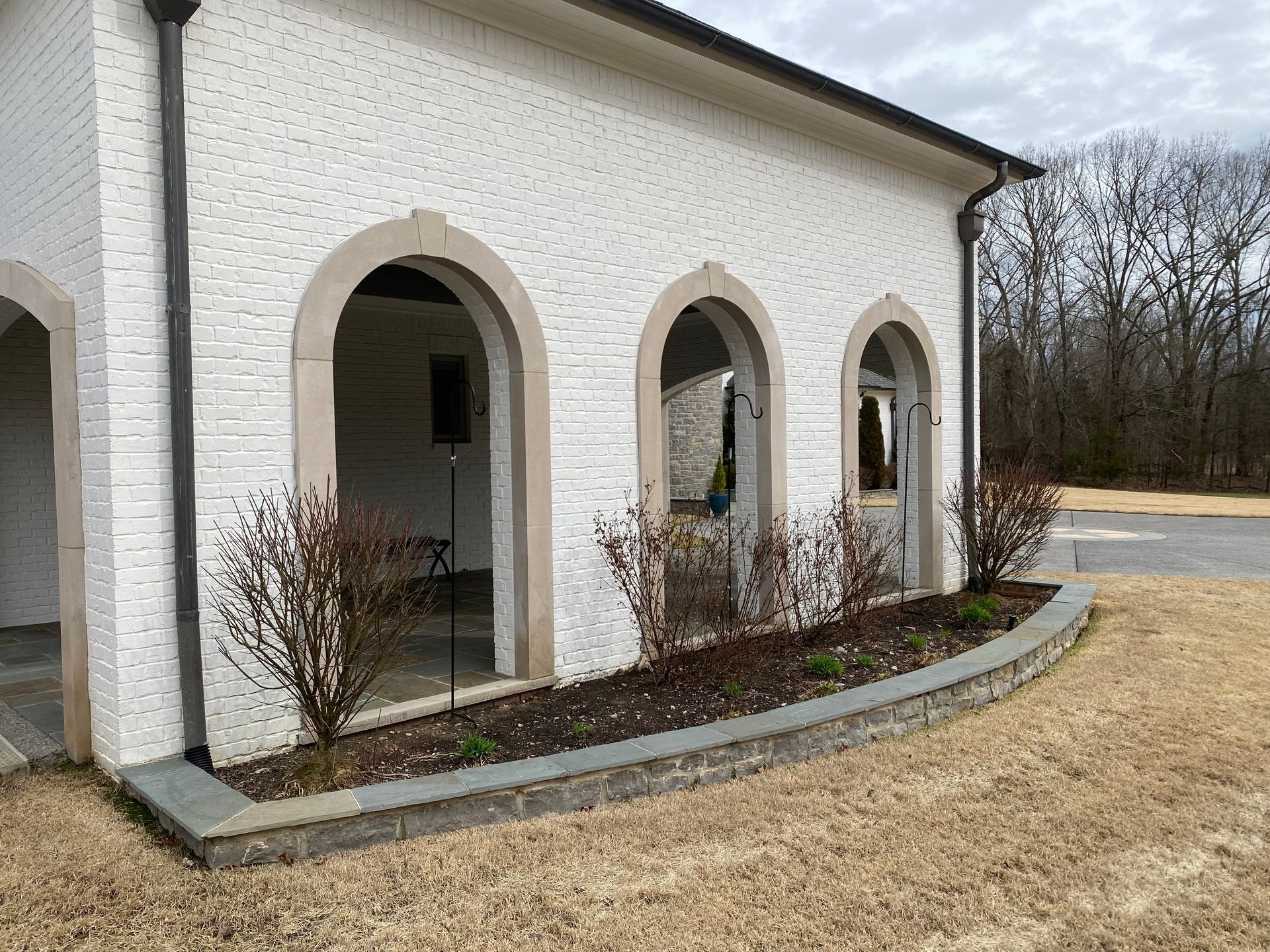 White brick building with arched openings, stone trim, and a low stone retaining wall with shrubs in front.