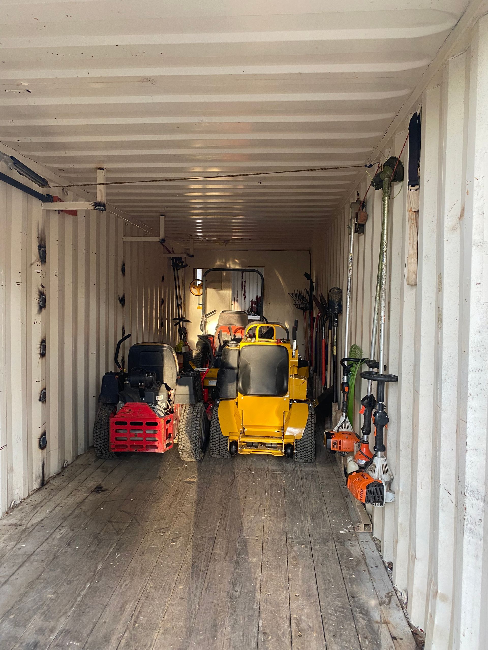 Inside a cargo container, a yellow construction machine and other equipment are stored, with tools mounted on the walls.