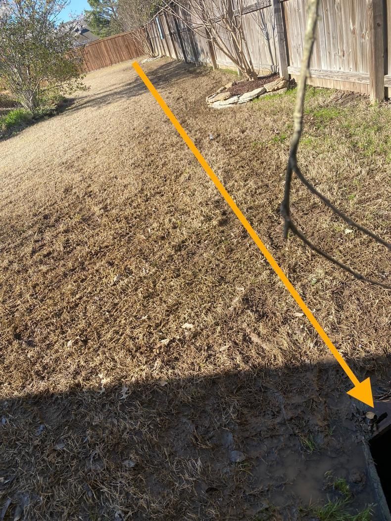 Overhead view of a backyard with brown grass, a path, and a fence. An orange arrow points diagonally across the yard toward a dark opening in the foreground.
