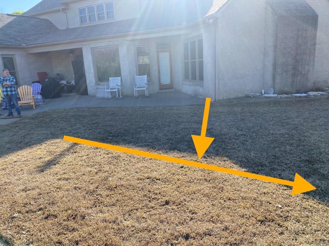 A sunny, low-angle view of dry grass in front of a house, with an arrow pointing to the right and another towards the back.
