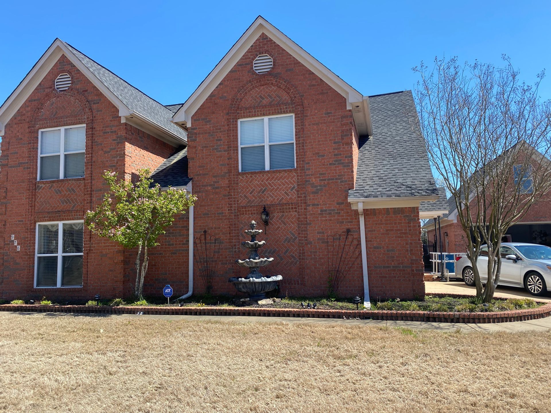 Red brick two-story house with a water fountain in front. Beige lawn, blue sky, and a car parked to the right.