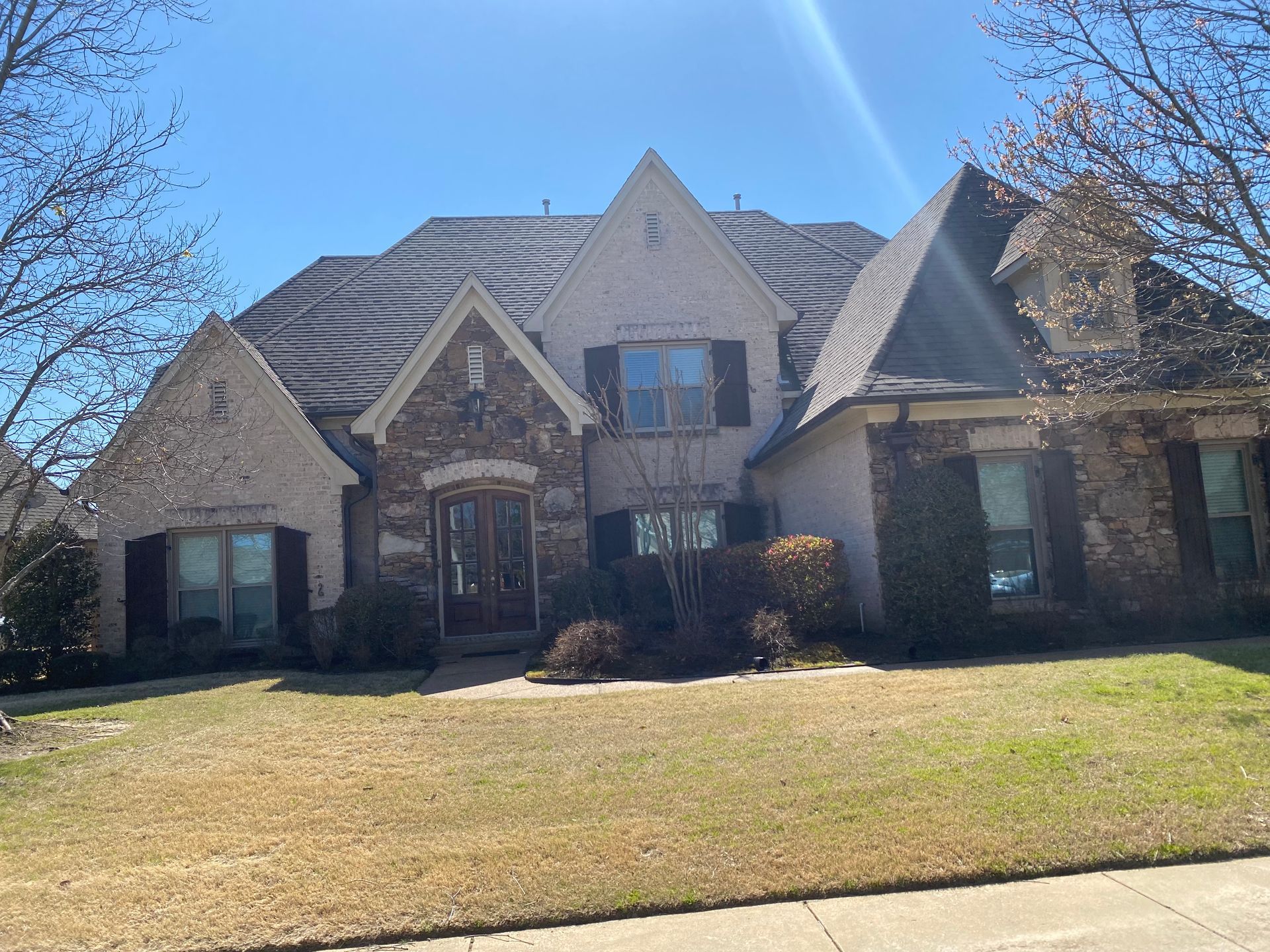 A two-story house with a light beige stucco facade, brown stone accents, and a brown shingle roof. The yard has brown grass and some bare trees.