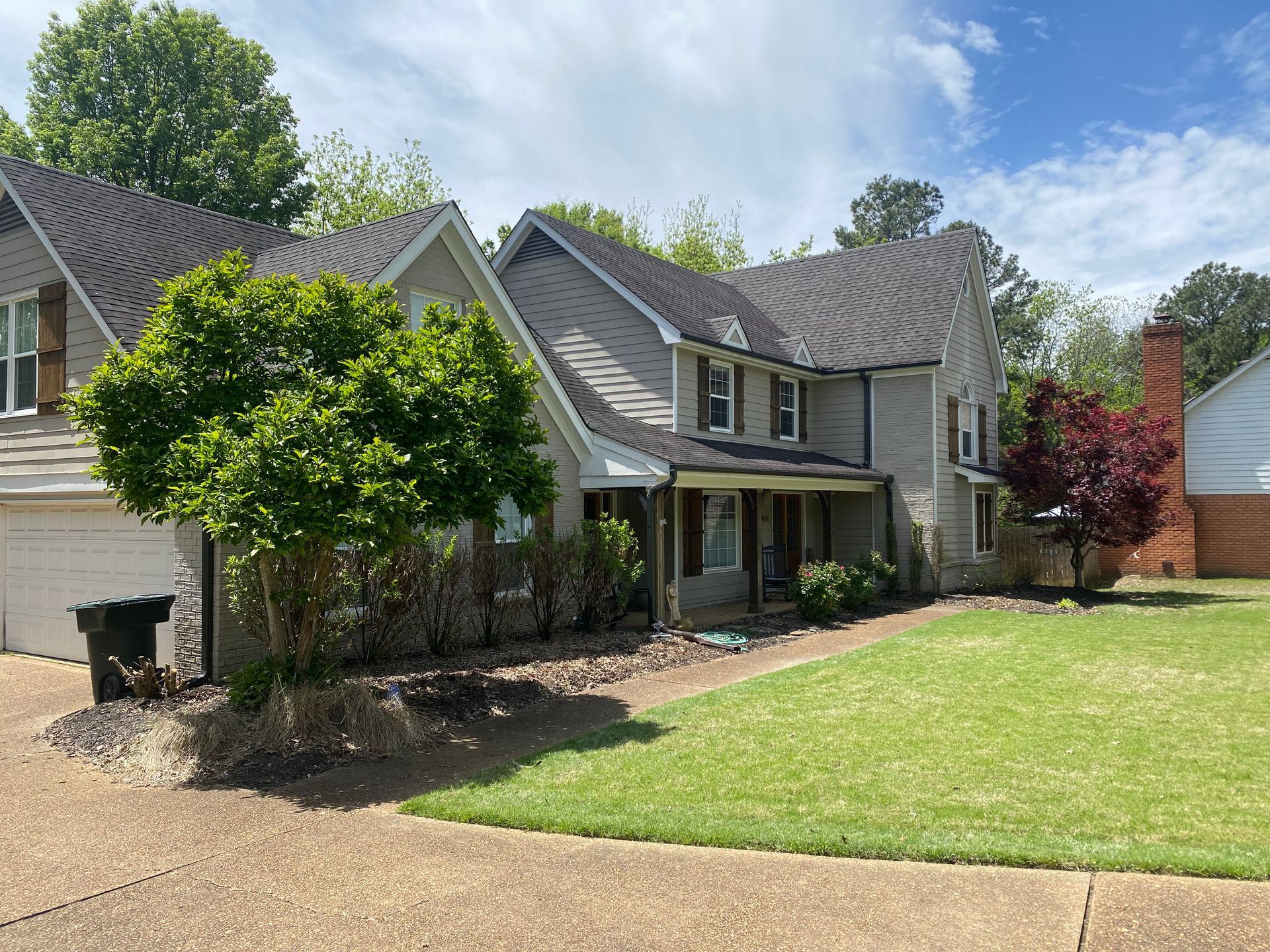 Two-story house with gray siding, black roof, and green lawn. A tree and bushes partially obscure the front.