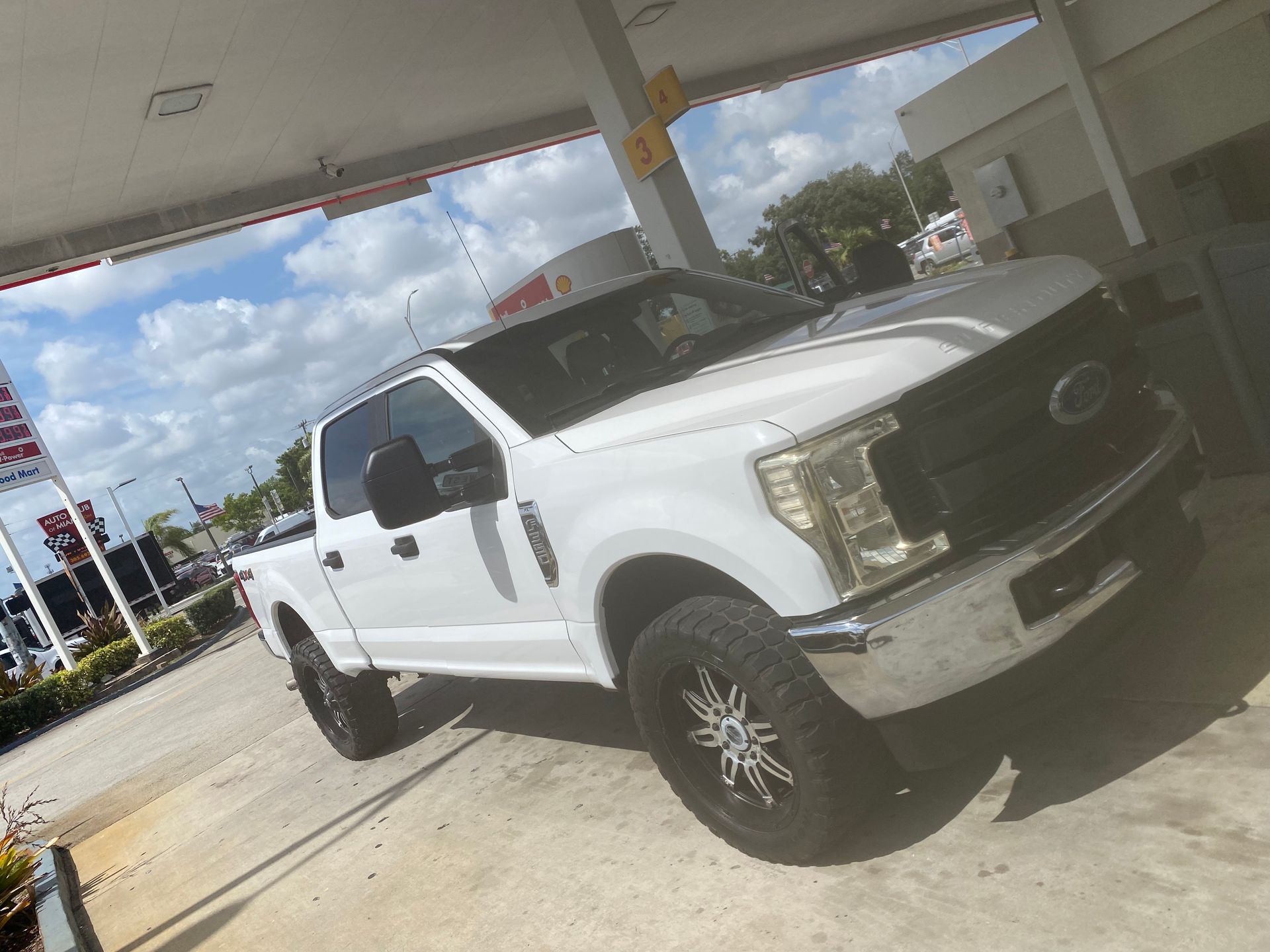 White Ford pickup truck parked at a Shell gas station on a sunny day. The truck has large black wheels.