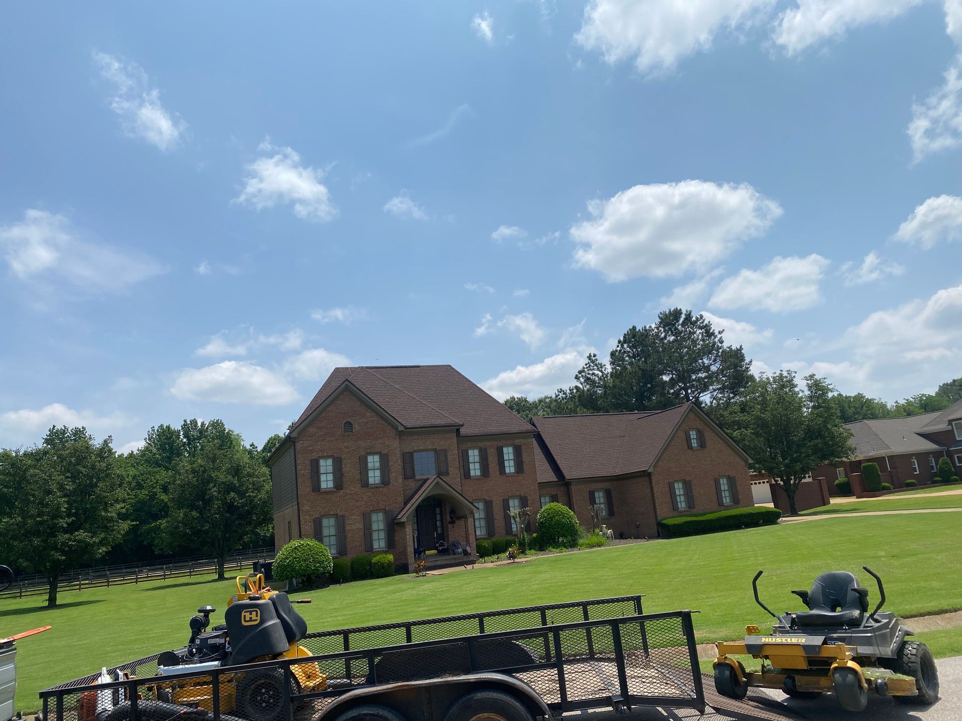 A brick house with a brown roof sits on a green lawn under a blue sky. A trailer with lawn equipment is in the foreground.