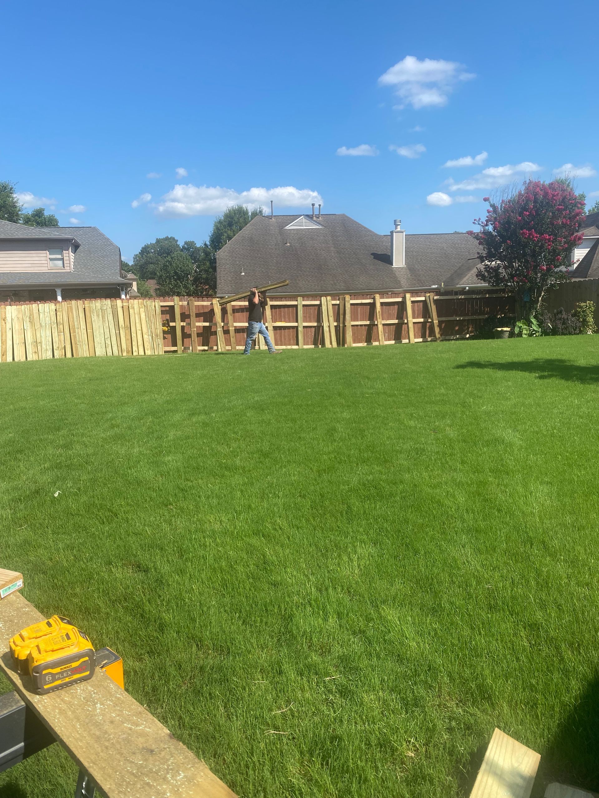 A person builds a wooden fence in a grassy backyard under a blue sky. Tools are visible in the foreground.